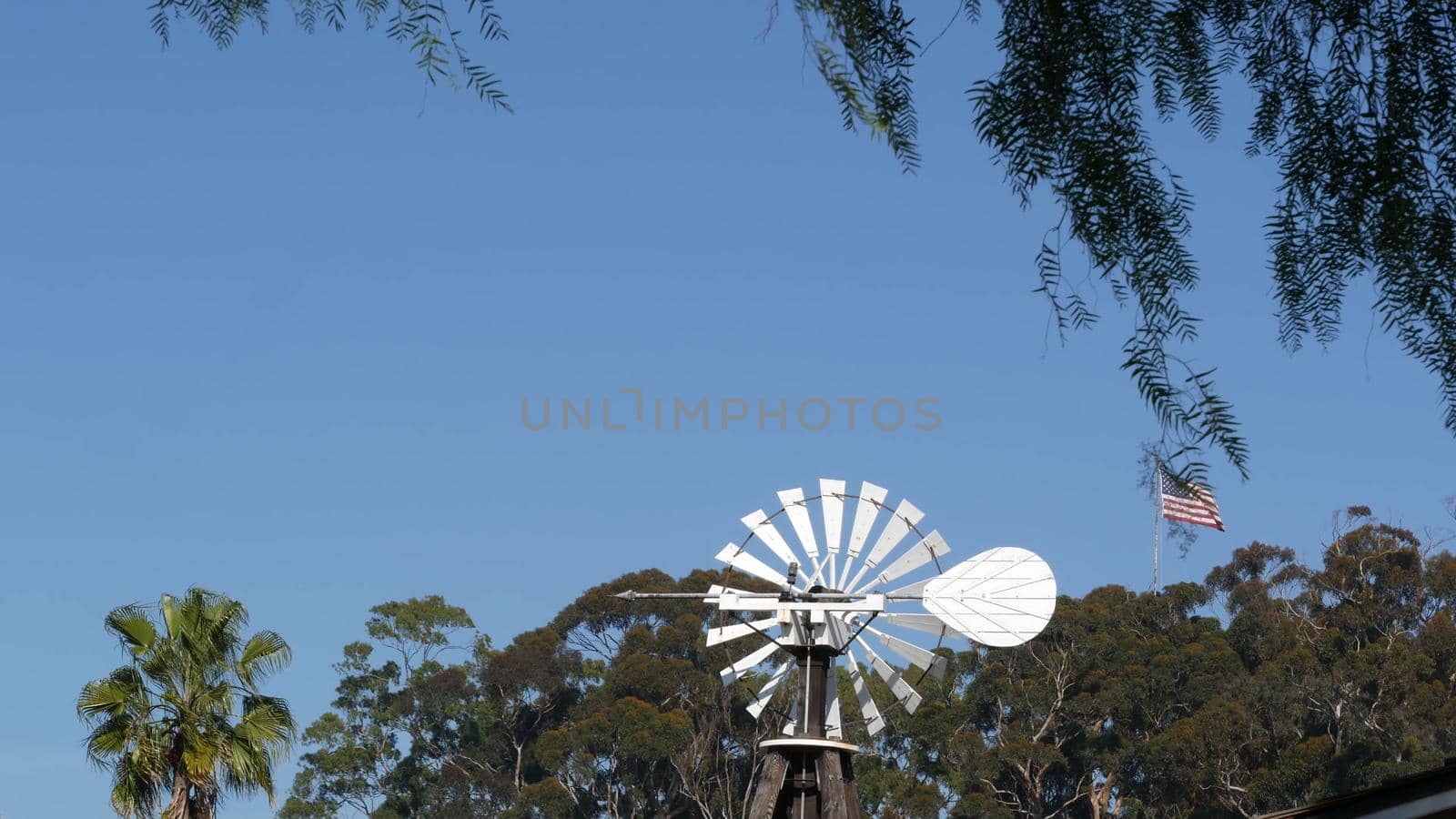 Classic retro windmill, bladed rotor and USA flag against blue sky. Vintage water pump wind turbine, power generator on livestock ranch or agricultural farm. Rural symbol of wild west, rustic suburb.