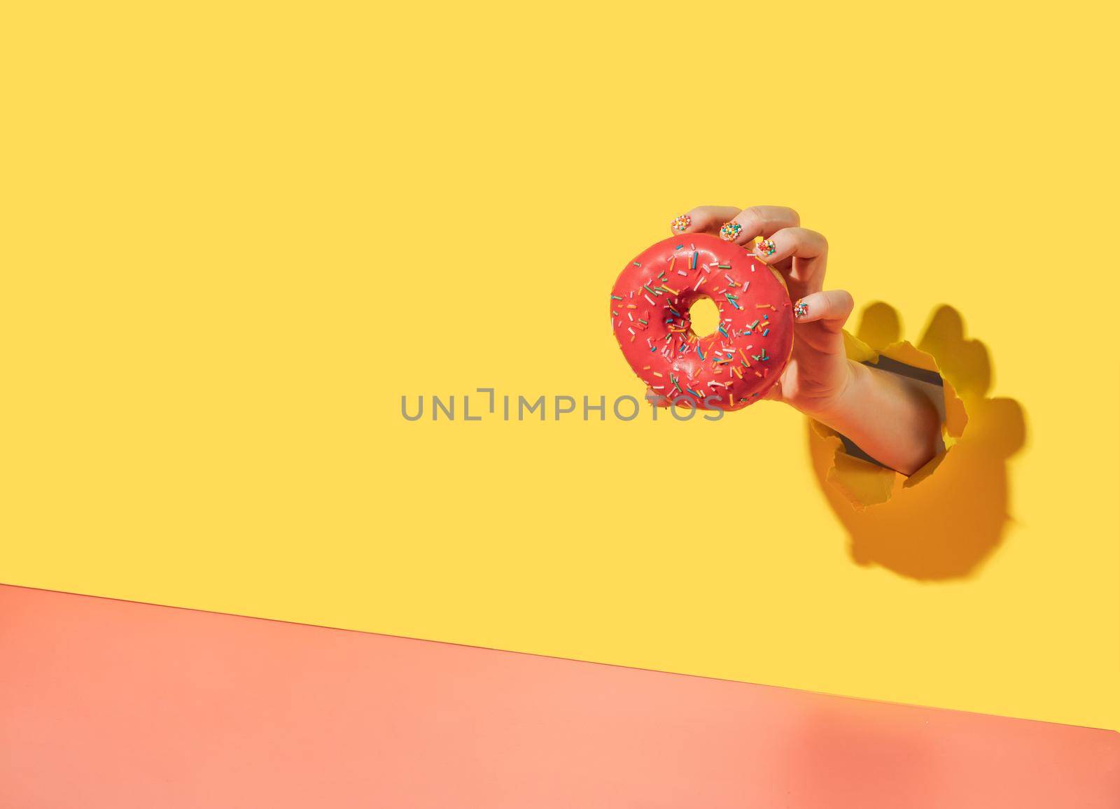 Donut in hand through torn bright yellow paper background. Donut delivery, diet, sugar concept. Female hand with sprinkle manicure holds glazed doughnut with sprinkle. Copy space. Creative still life