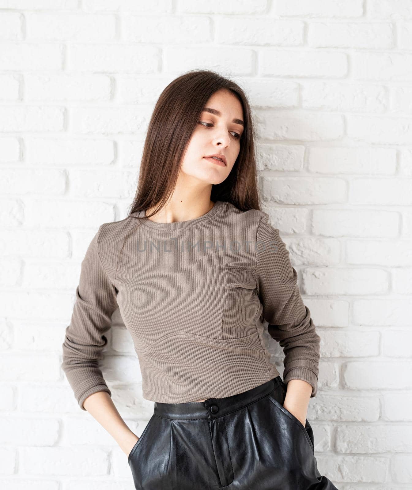 Three quarters length portrait of beautiful smiling brunette woman with long hair wearing brown shirt and black leather shorts, on white brick wall background, hands in pockets