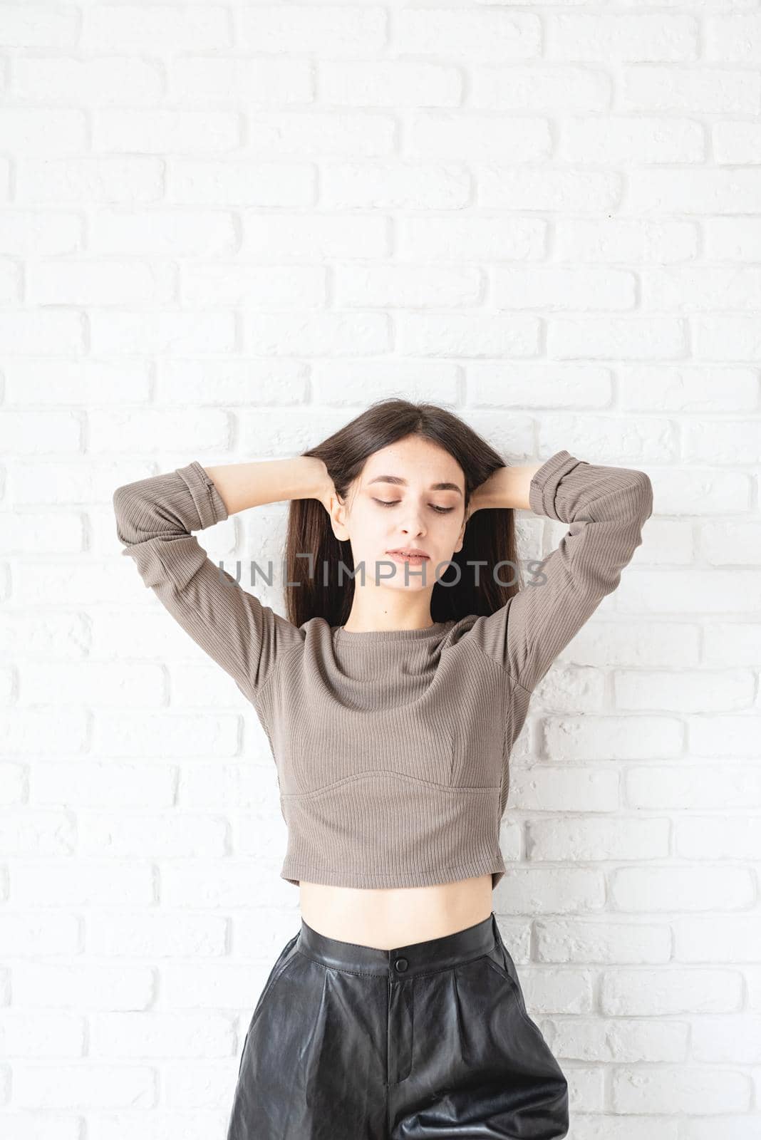 Three quarters length portrait of beautiful smiling brunette woman with long hair wearing brown shirt and black leather shorts, on white brick wall background