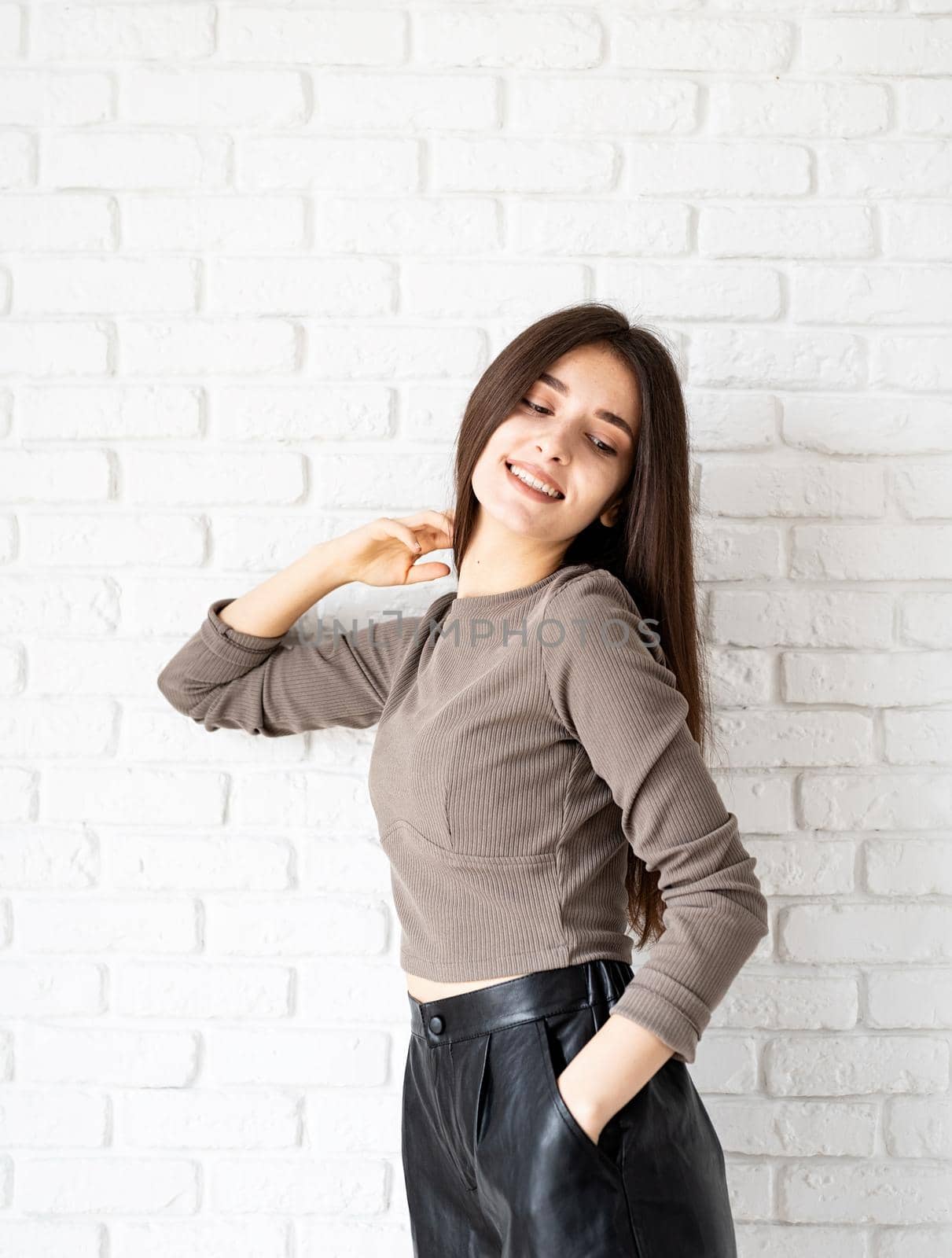 Three quarters length portrait of beautiful smiling brunette woman with long hair wearing brown shirt and black leather shorts, on white brick wall background