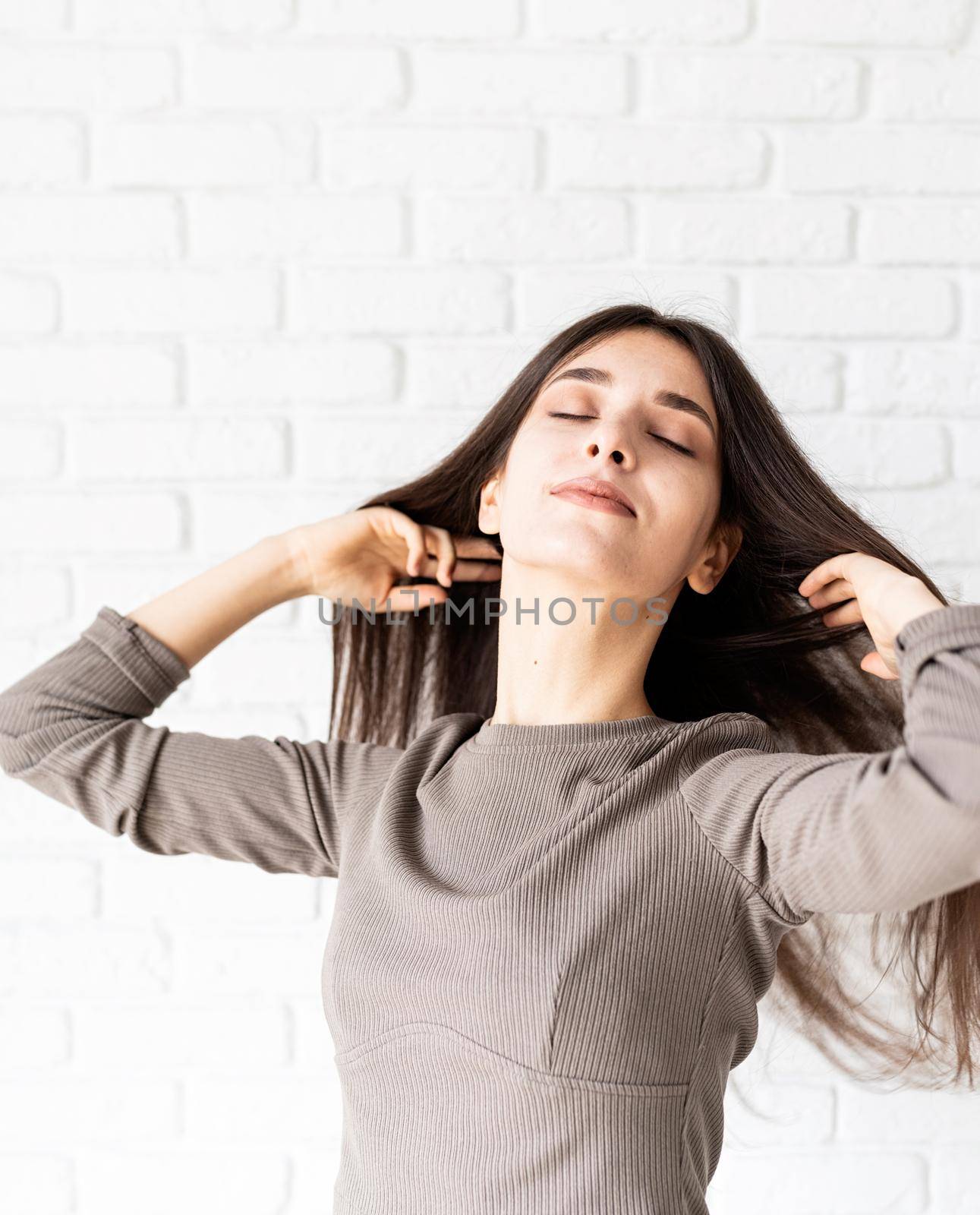 Three quarters length portrait of beautiful smiling brunette woman with long hair wearing brown shirt and black leather shorts, on white brick wall background with eyes closed