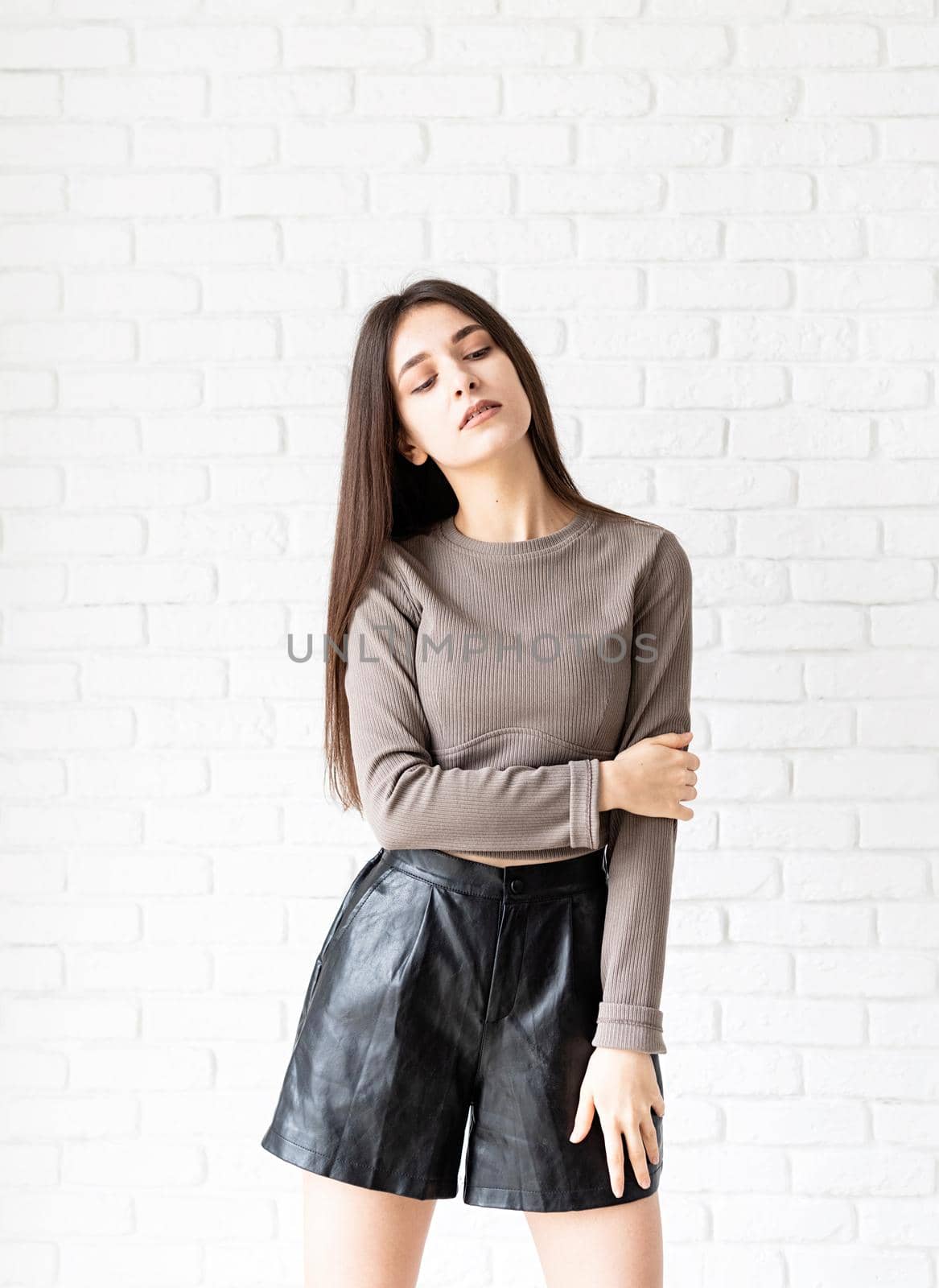 Three quarters length portrait of beautiful smiling brunette woman with long hair wearing brown shirt and black leather shorts, on white brick wall background