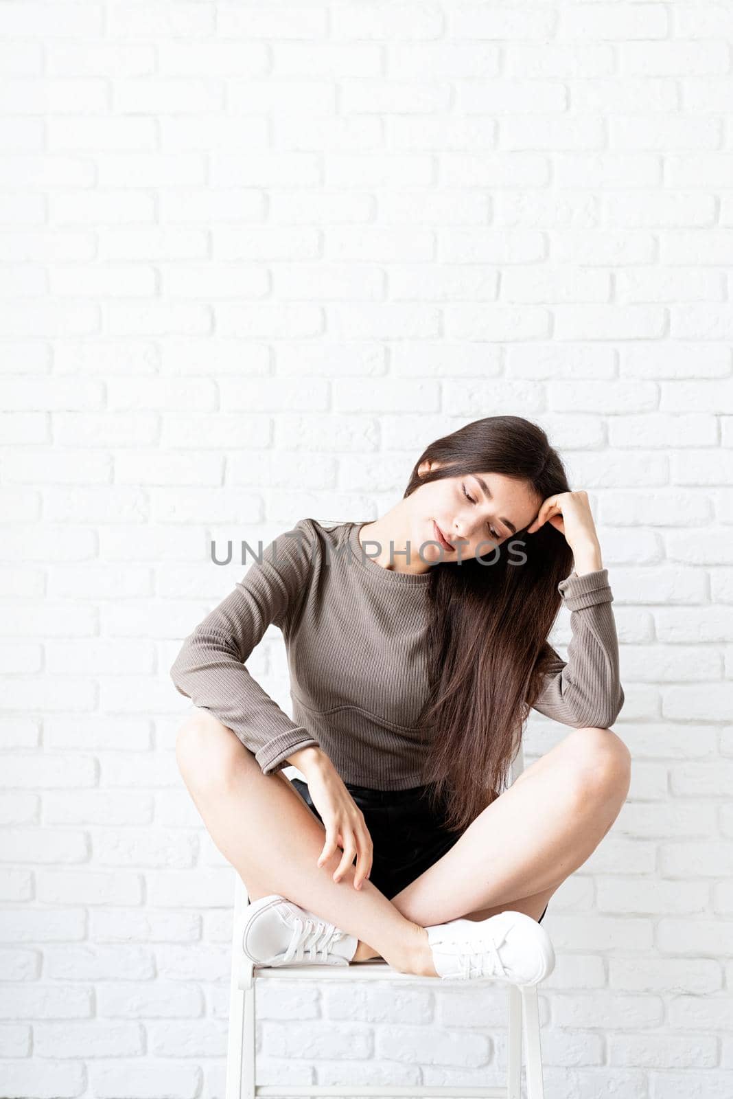 portrait of a beautiful brunette woman with long hair sitting on white brick wall background, thinking by Desperada