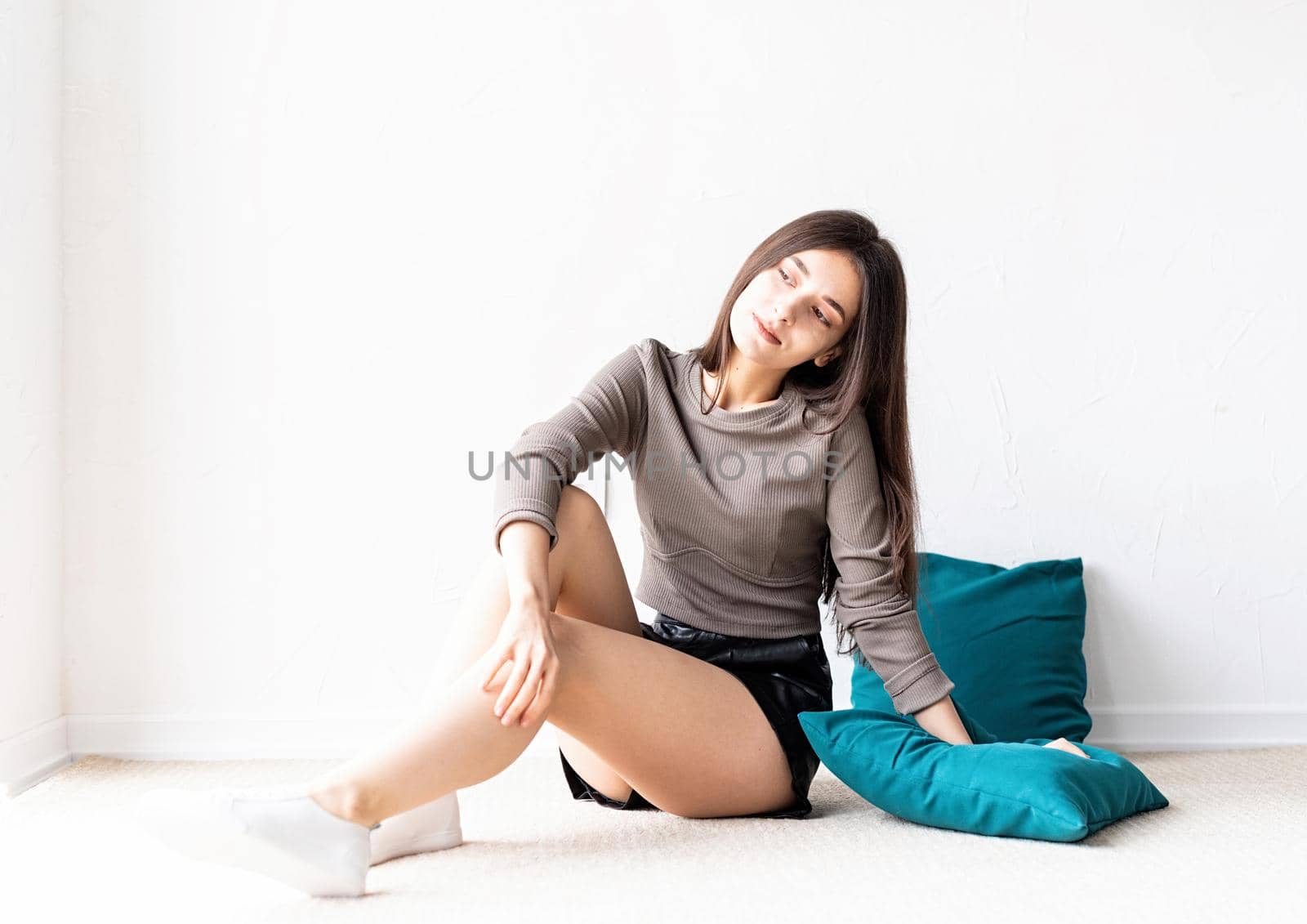 Beautiful young woman in casual clothes sitting on the floor with pillows smiling looking away