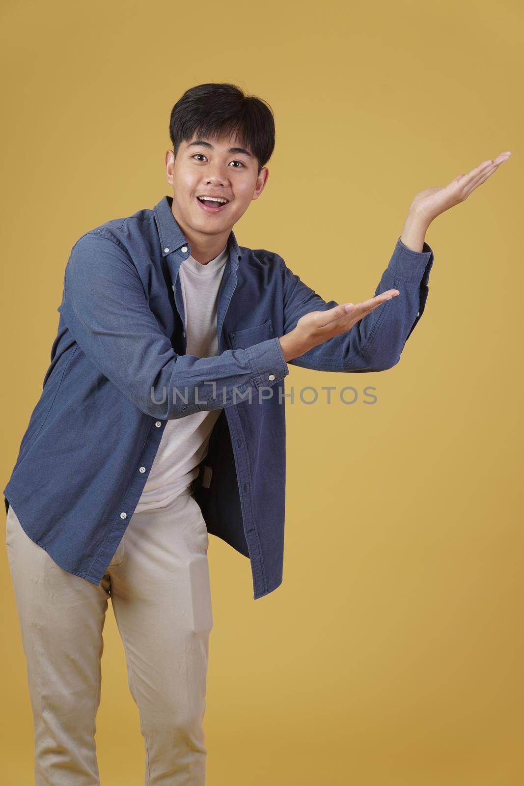portrait of happy handsome young asian man dressed casually presenting with open palms gesture at copyspace. yellow studio background