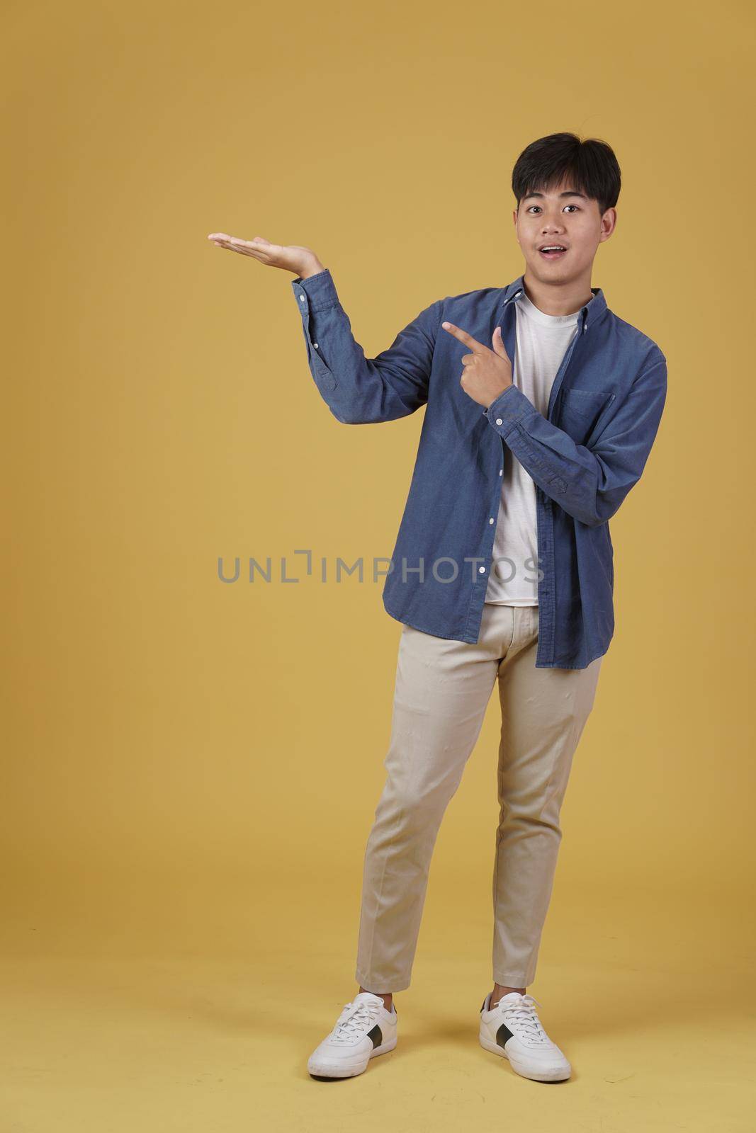 portrait of happy smiling young asian man dressed casually pointing finger to empty open hand isolated on yellow background