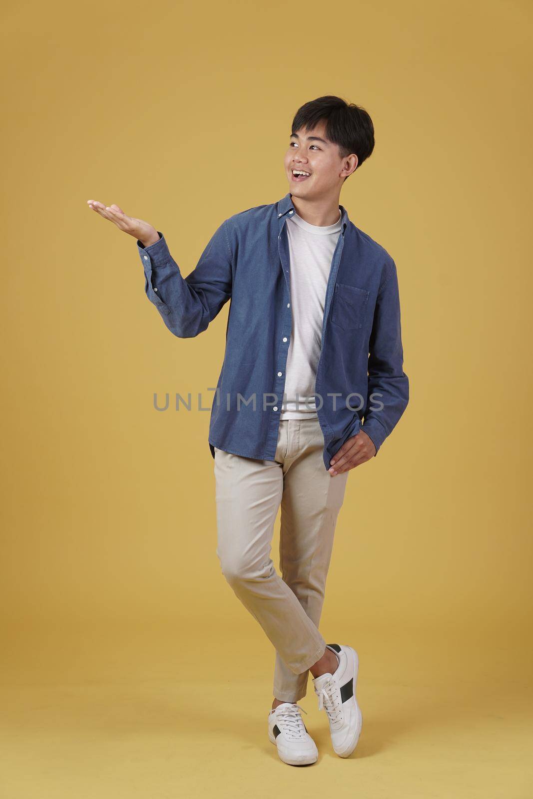 portrait of excited happy handsome young asian man dressed casually smiling with open hand gesture isolated on yellow studio background