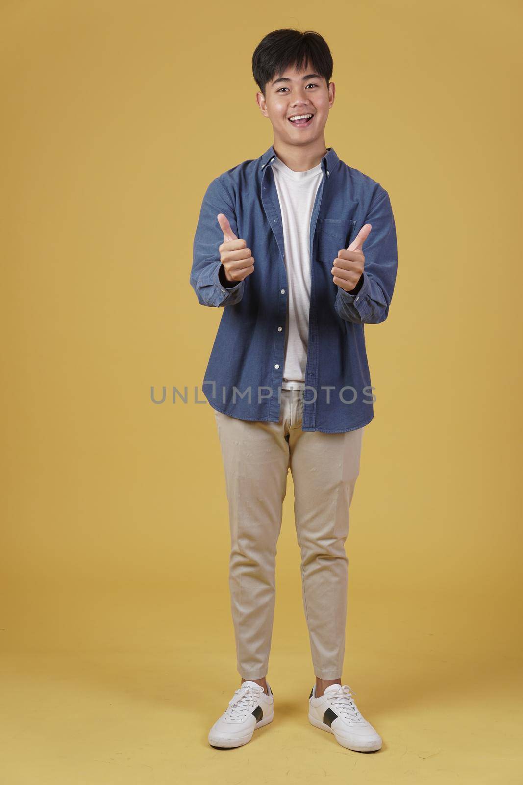 portrait of smiling positive young asian man dressed casually with thumb up gesture approving expression isolated on yellow studio background