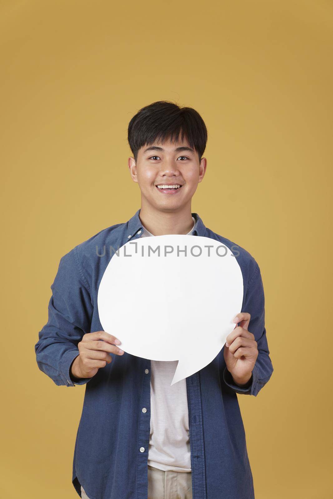 portrait of smiling happy cheerful young asian man dressed casually with empty speech bubble isolated on yellow studio background