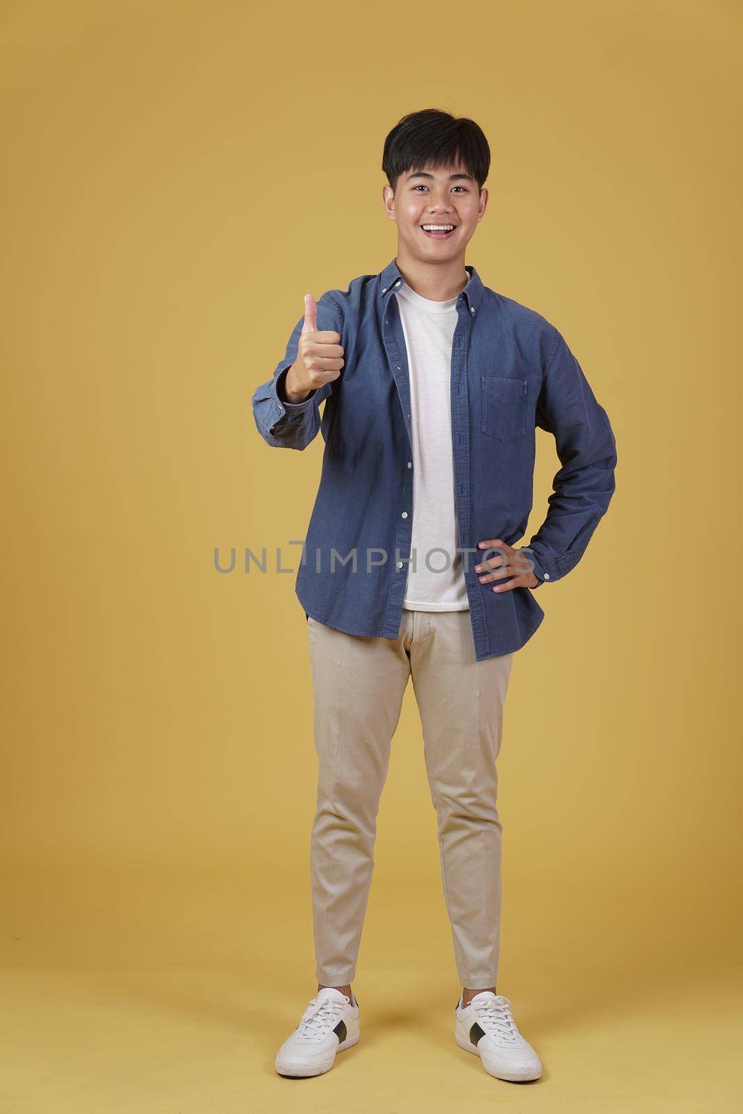 portrait of smiling positive young asian man dressed casually with thumb up gesture approving expression isolated on yellow studio background