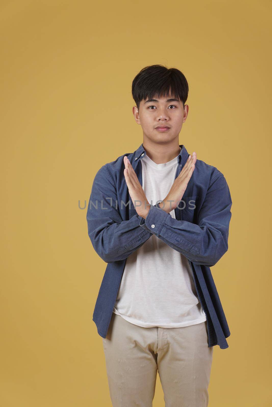 portrait of handsome young asian man dressed casually crossing arms in negative rejection or no expression isolated on yellow studio background