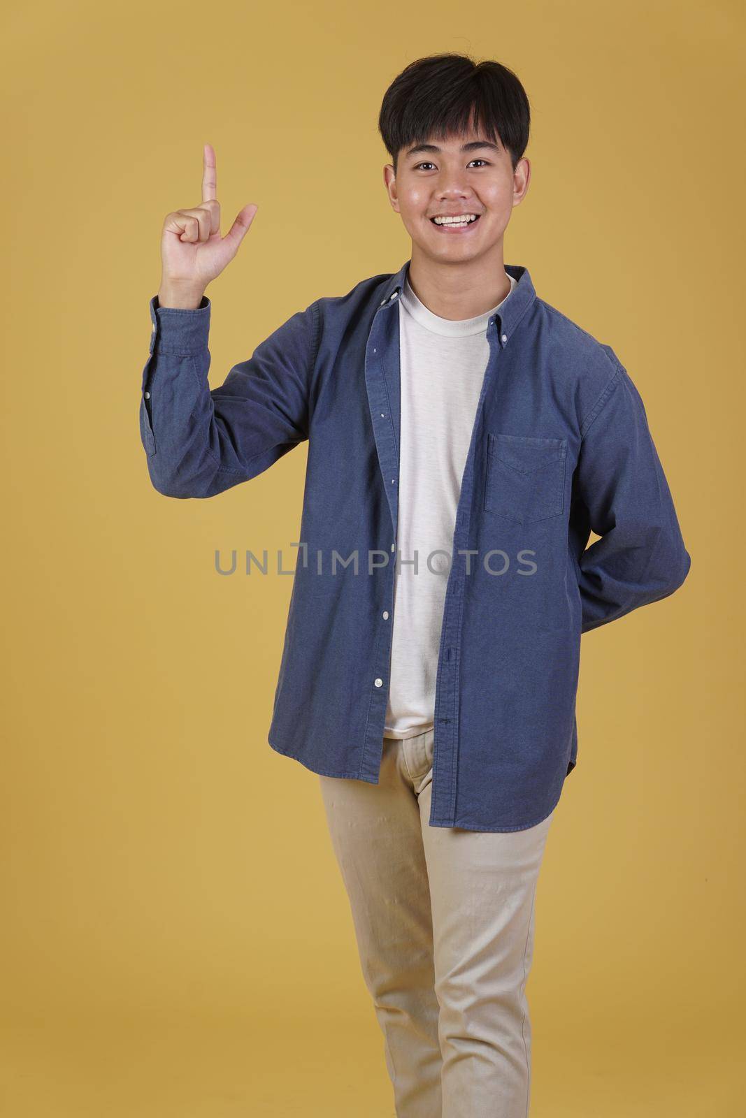 portrait of happy young asian man dressed casually pointing finger up at copyspace. yellow studio background