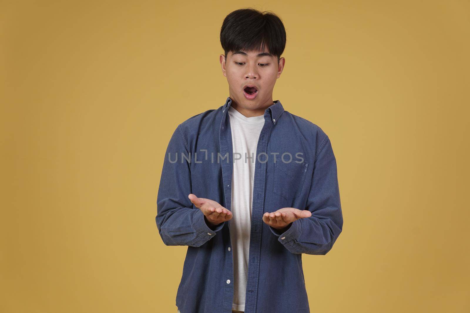 portrait of surprised excited young asian man dressed casually holding something on open palms isolated on yellow studio background