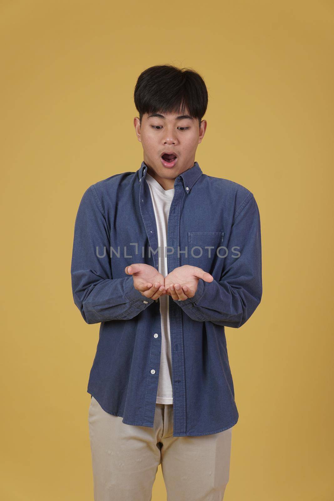 portrait of surprised excited young asian man dressed casually holding something on open palms isolated on yellow studio background