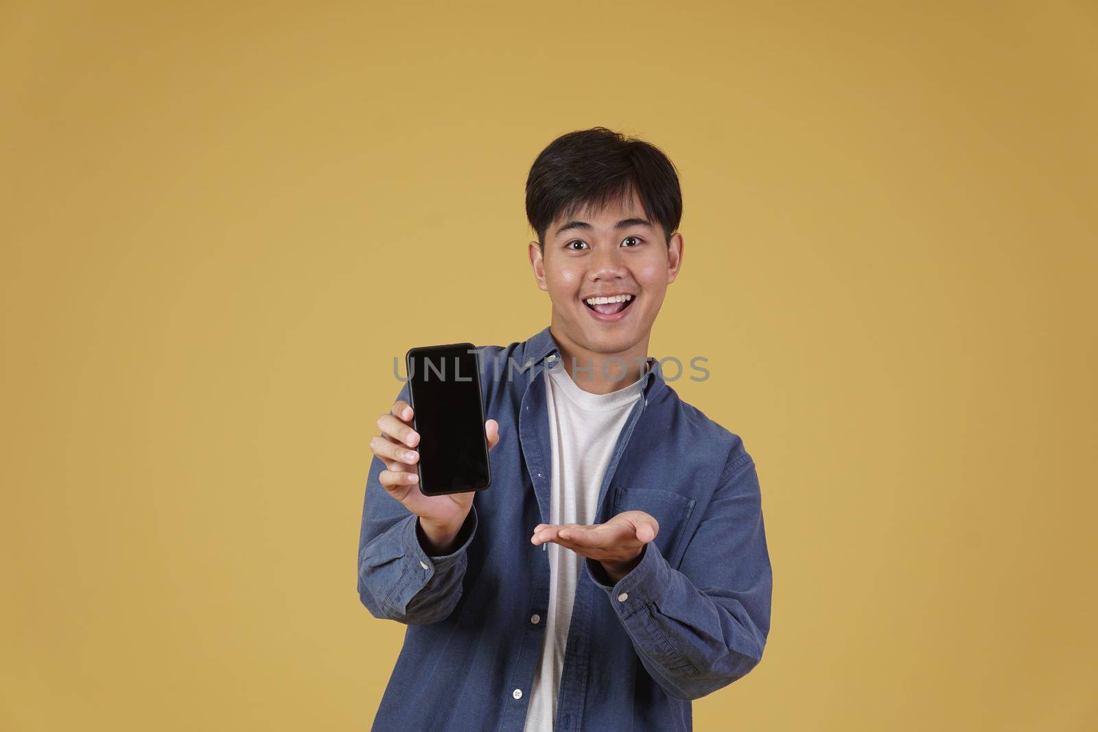 portrait of happy young asian man dressed casually smiling showing smartphone blank white screen isolated on yellow studio background