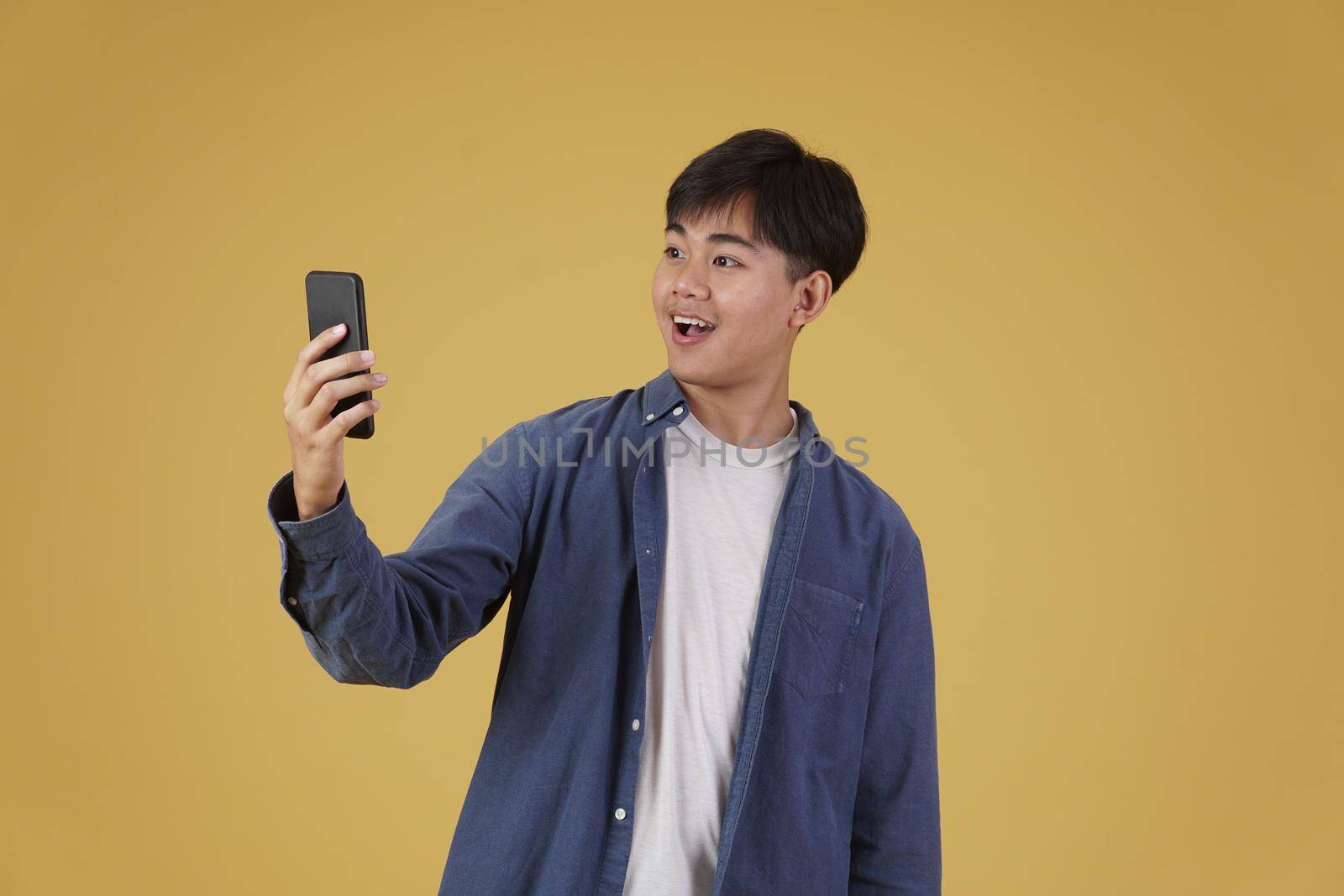 portrait of happy surprised excited young asian man dressed casually looking at smartphone isolated on yellow studio background