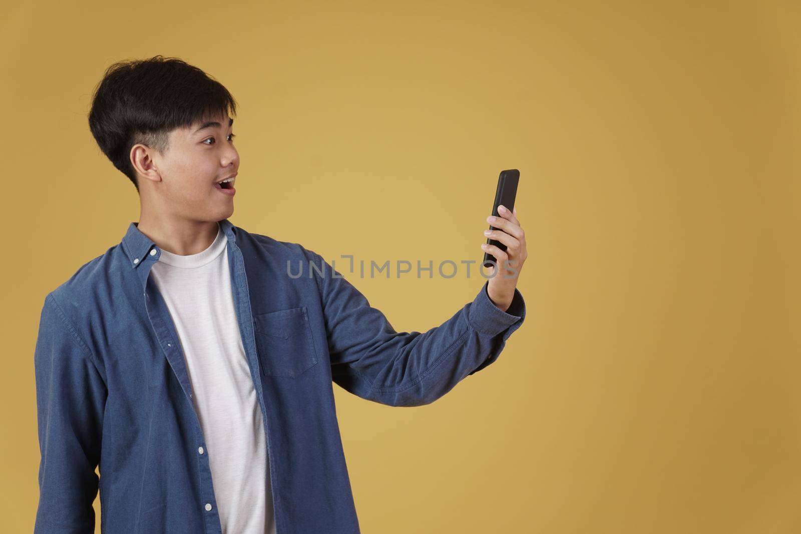 portrait of happy surprised excited young asian man dressed casually looking at smartphone isolated on yellow studio background