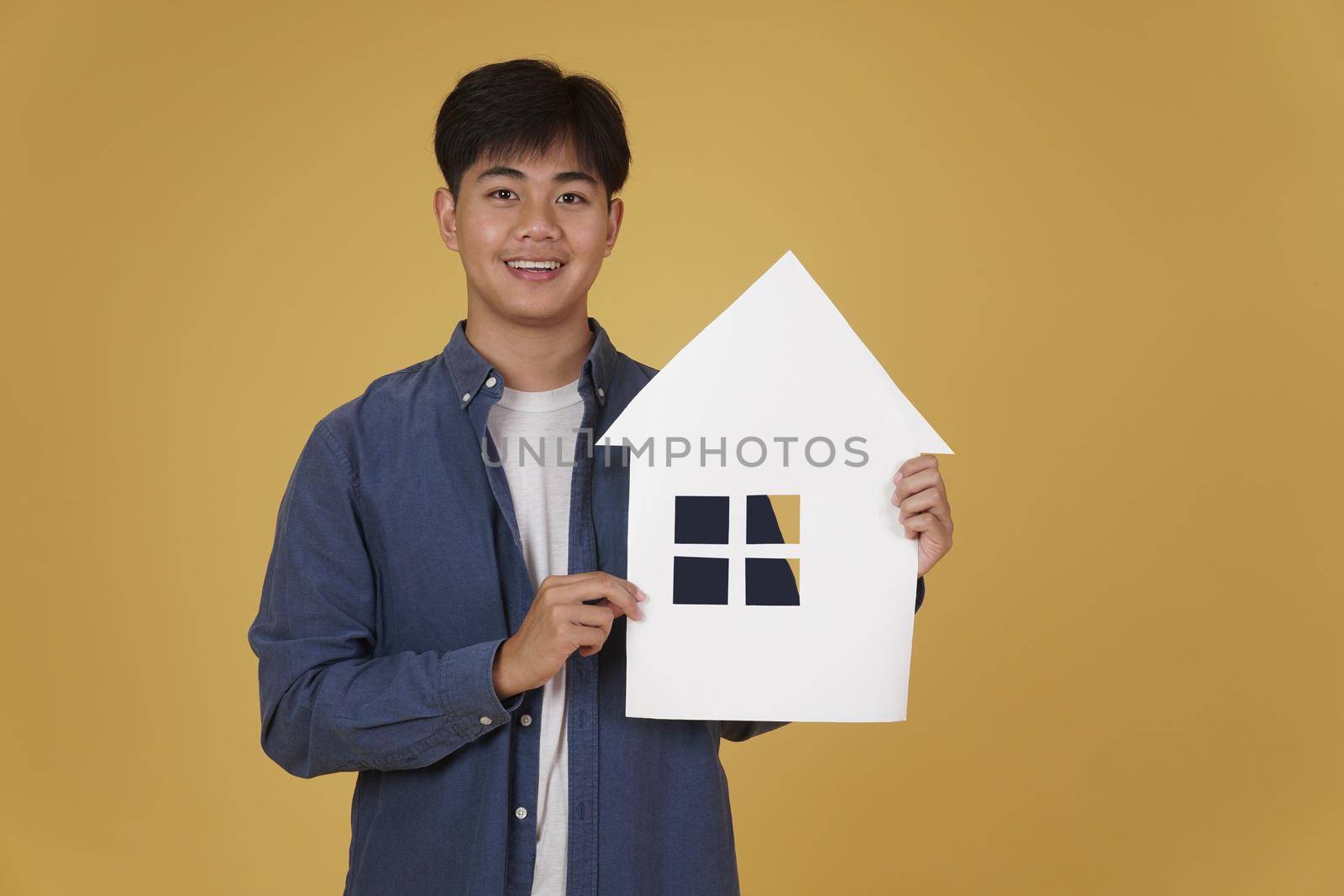 portrait of smiling happy cheerful young asian man dressed casually with home house paper cutout isolated on yellow studio background. real estate purchase concept