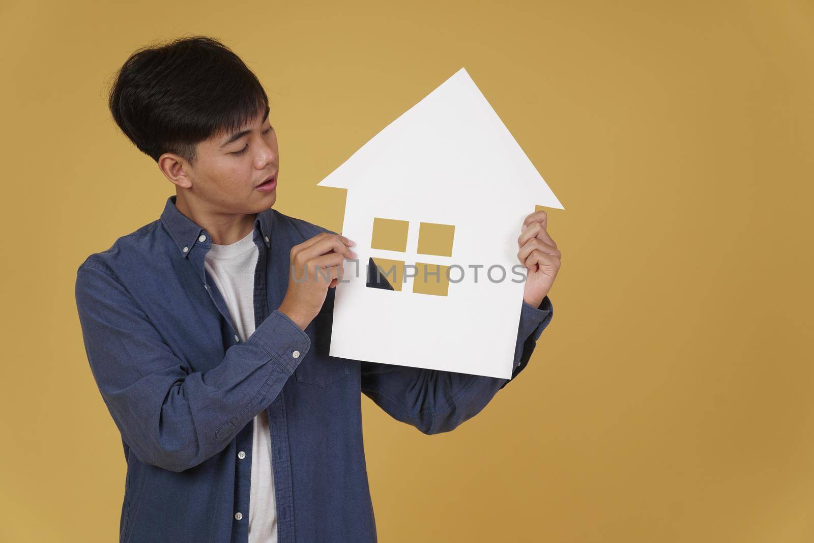 portrait of smiling happy cheerful young asian man dressed casually with home house paper cutout isolated on yellow studio background. real estate purchase concept