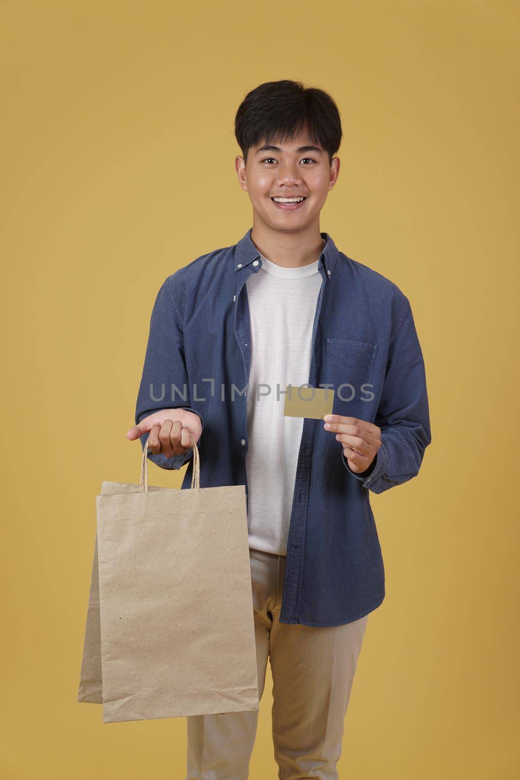 portrait of happy young asian man dressed casually holding shopping bags and credit card isolated on yellow studio background