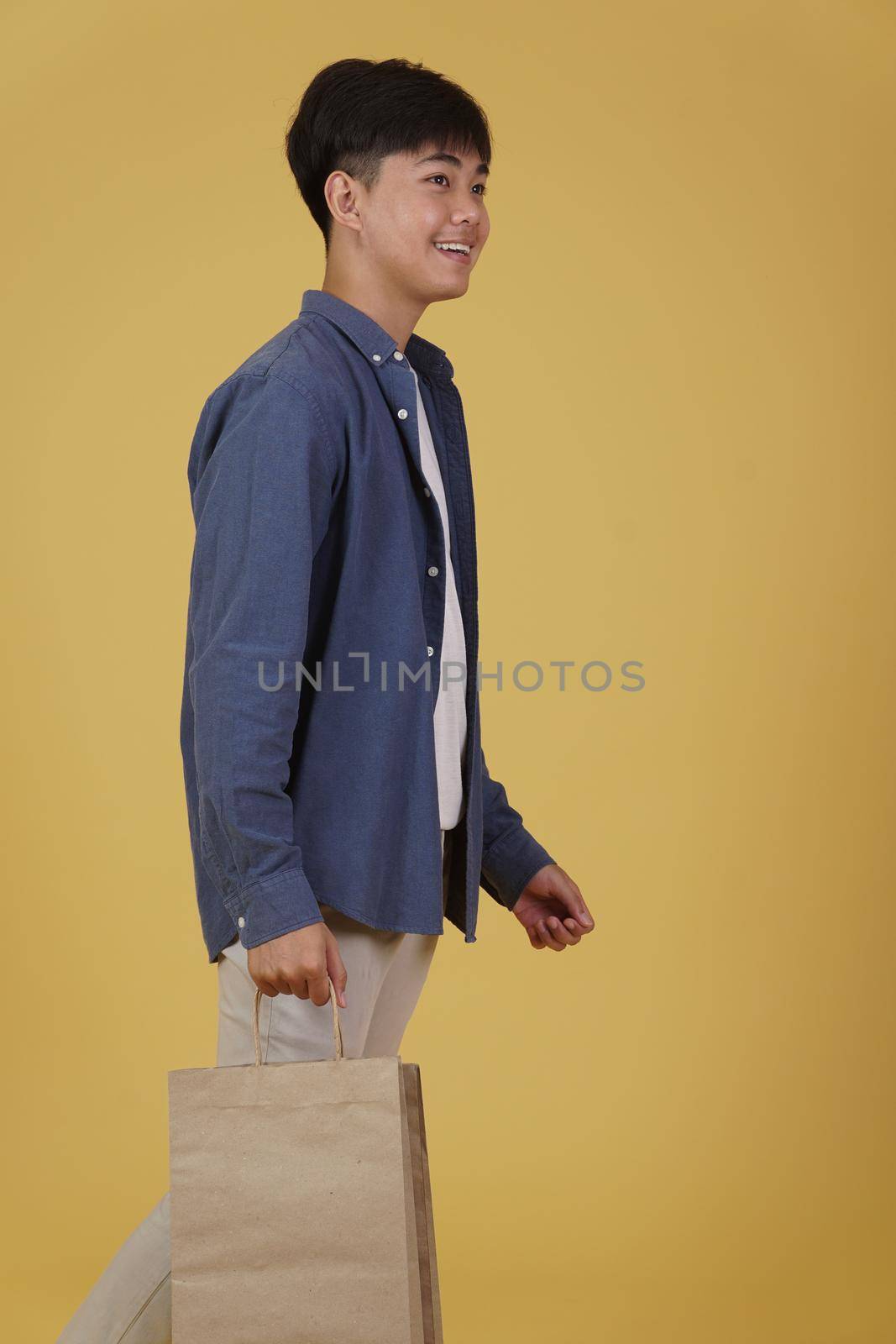 portrait of happy young asian man dressed casually holding shopping bags isolated on yellow studio background