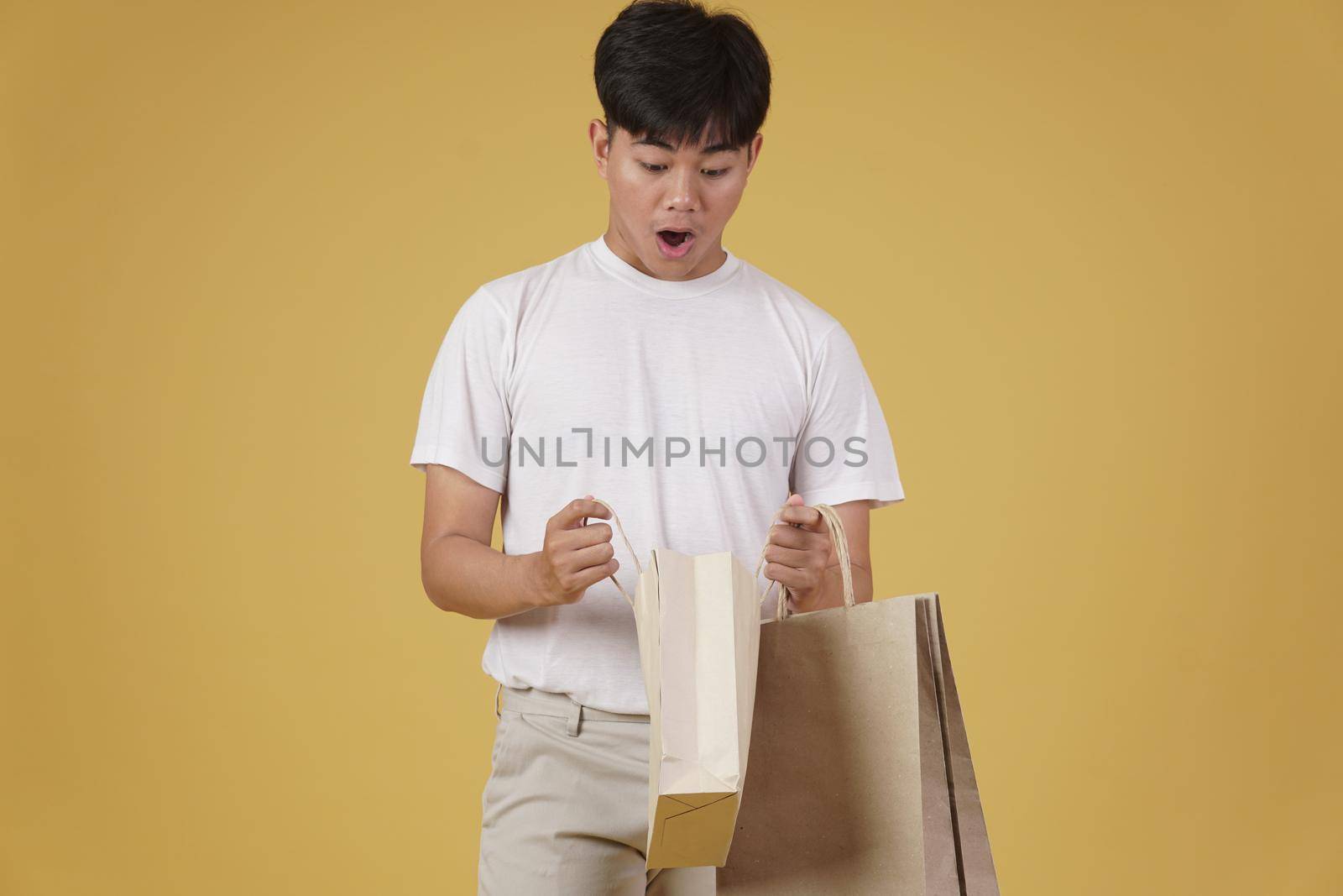 portrait of surprised excited young asian man dressed casually opening shopping bags isolated on yellow studio background