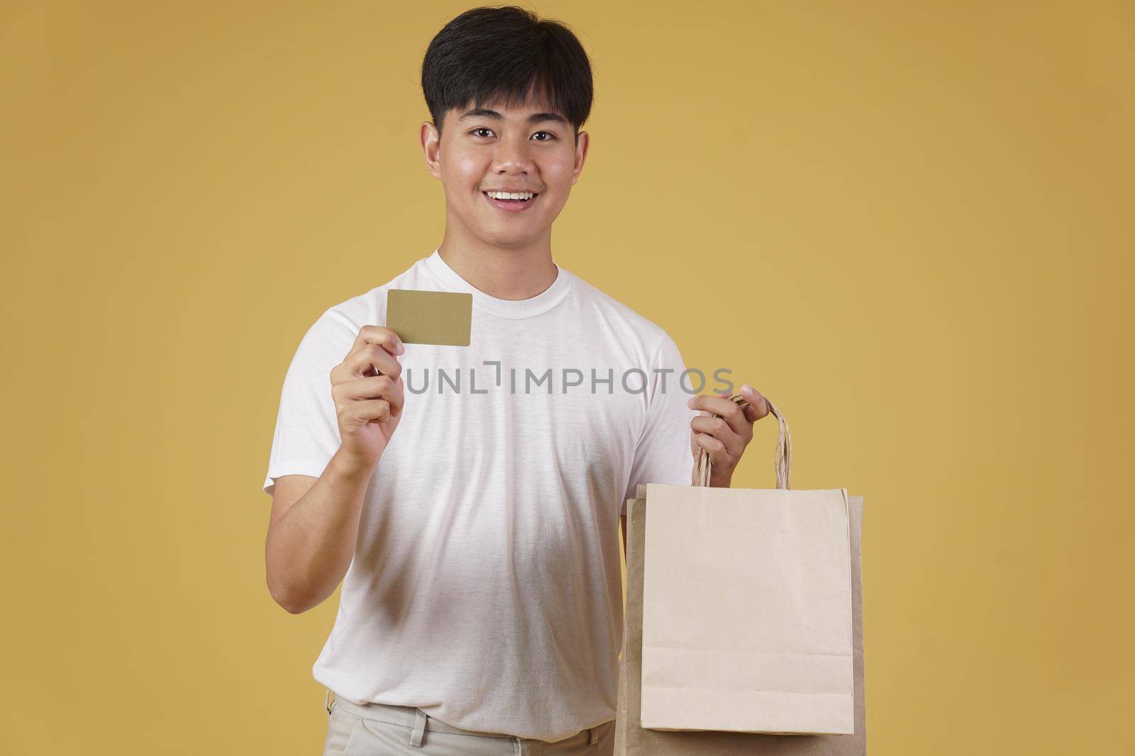 portrait of happy young asian man dressed casually holding shopping bags and credit card isolated on yellow studio background