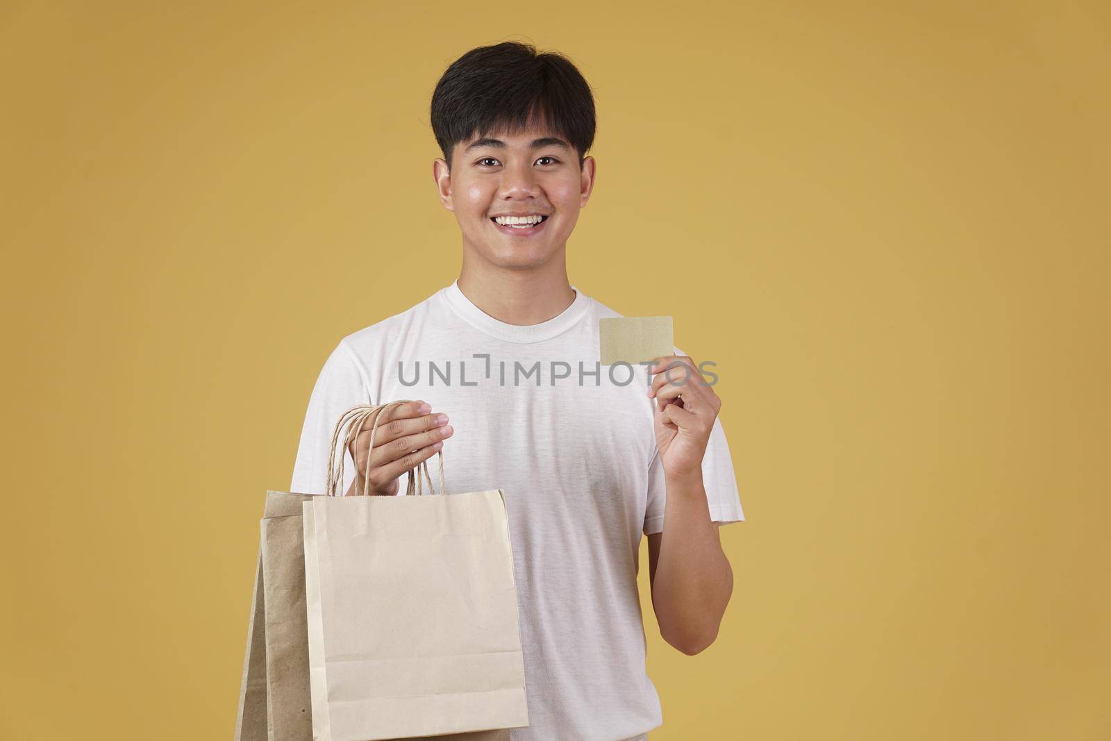portrait of happy young asian man dressed casually holding shopping bags and credit card isolated on yellow studio background