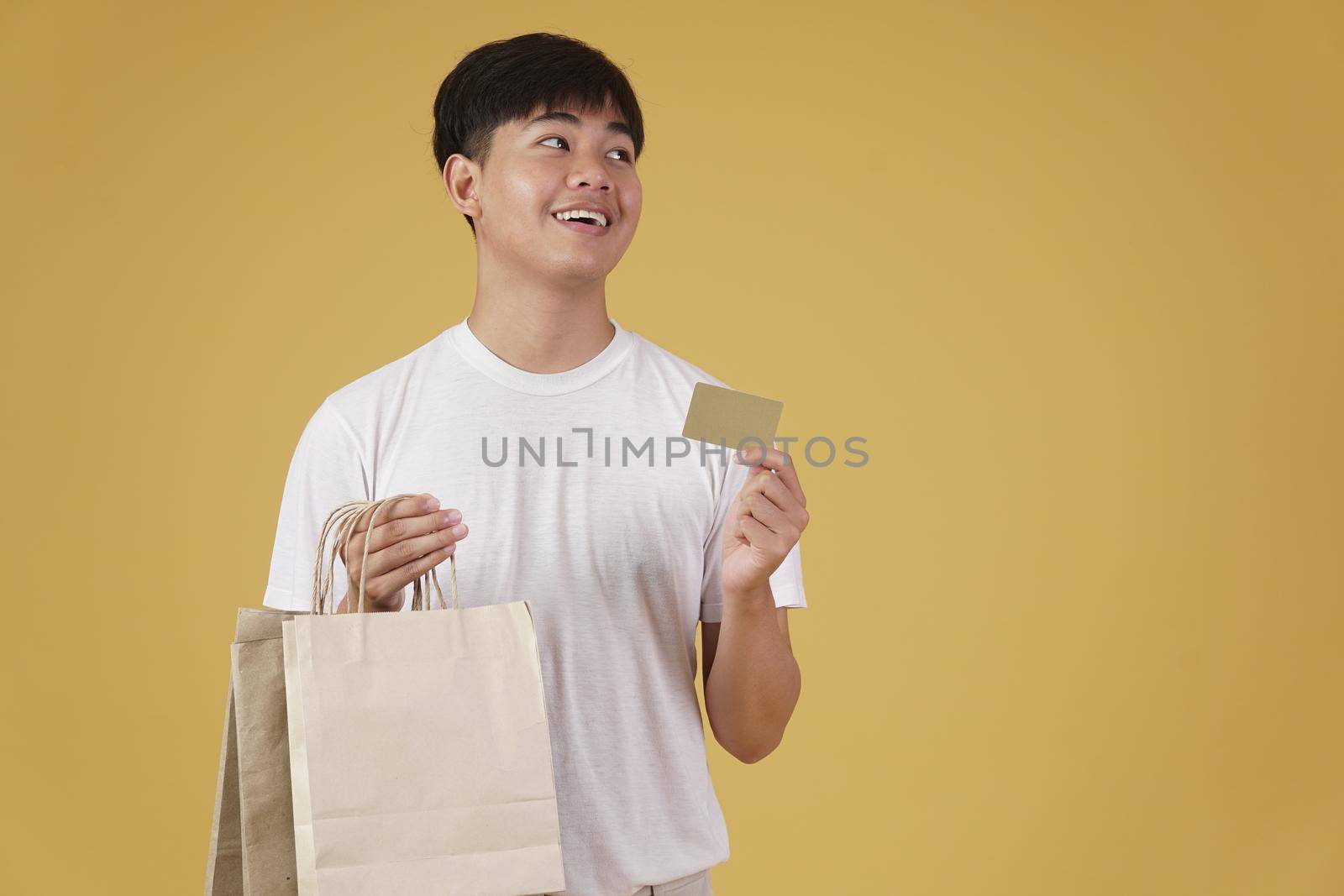 portrait of happy young asian man dressed casually holding shopping bags and credit card isolated on yellow studio background