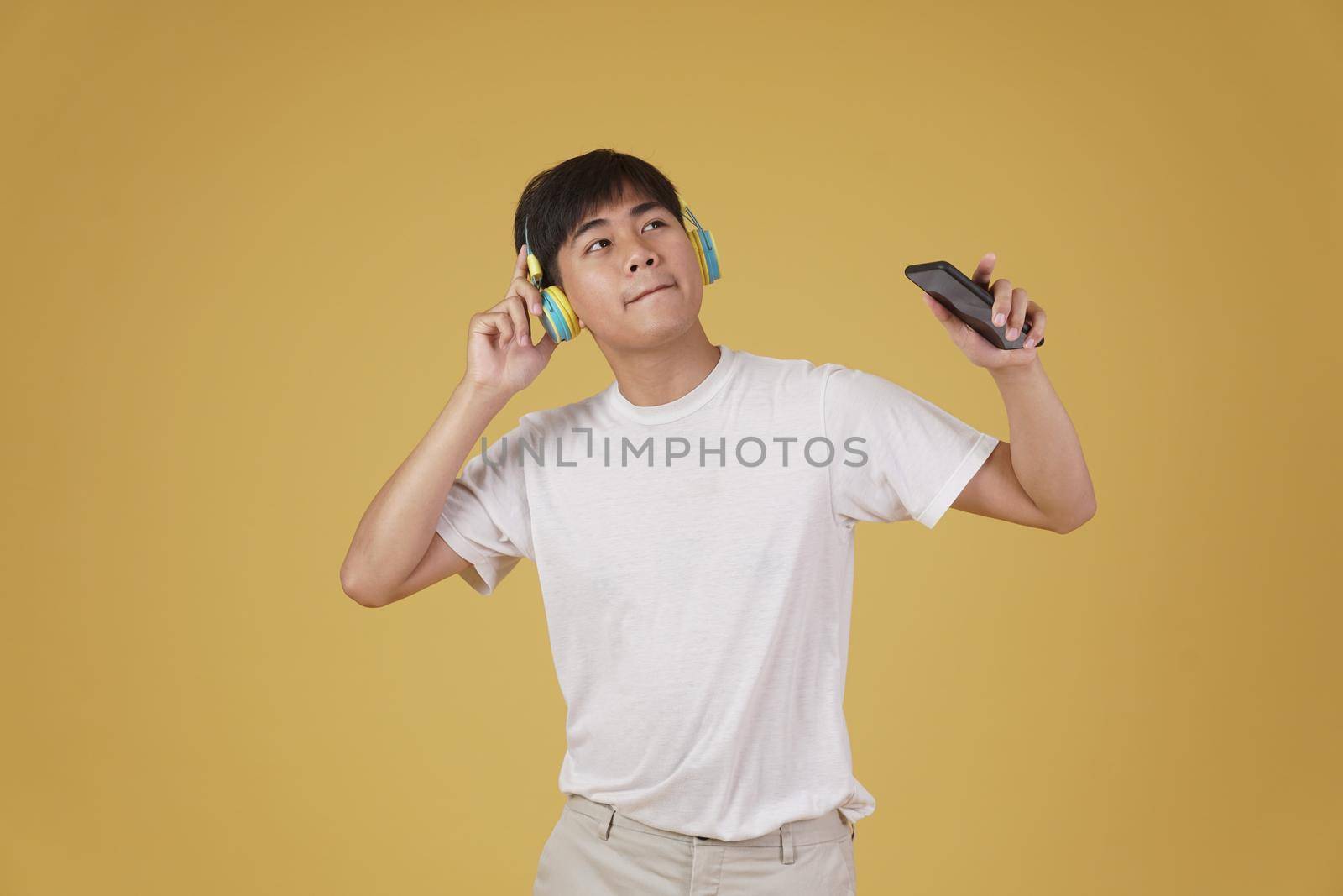 portrait of happy joyful cheerful young asian man wearing headphones listening to music on smartphone and dancing isolated on yellow studio background