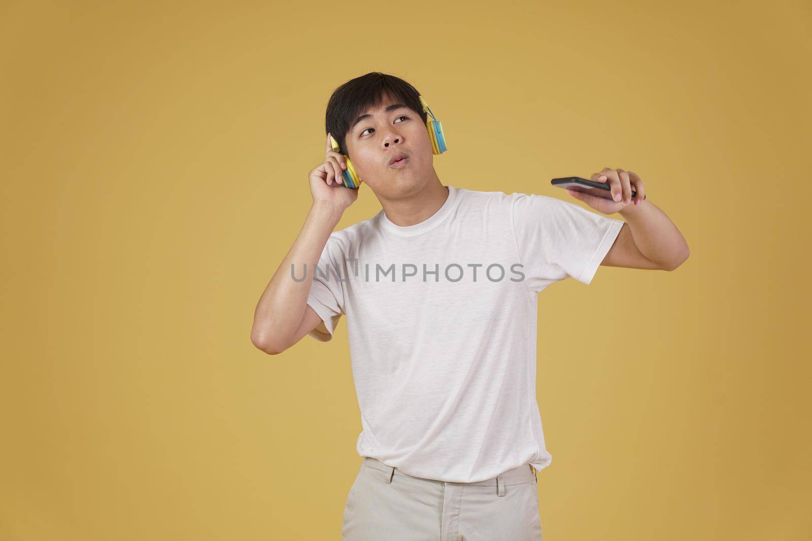 portrait of happy joyful cheerful young asian man wearing headphones listening to music on smartphone and dancing isolated on yellow studio background