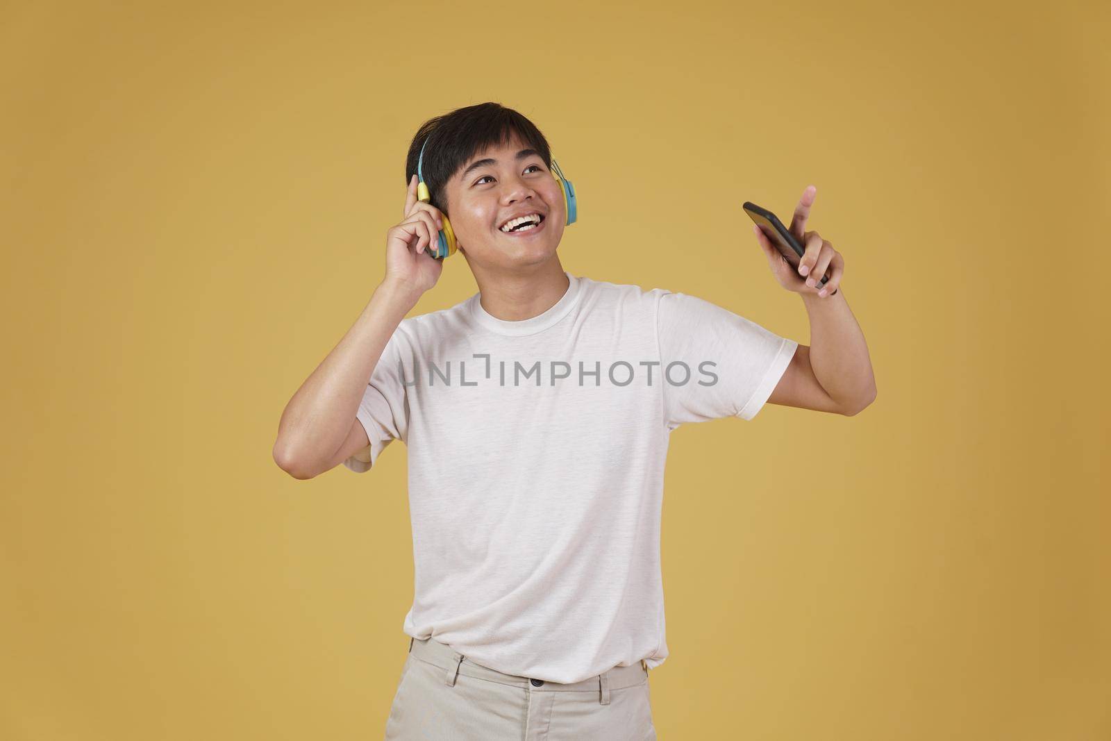 portrait of happy joyful cheerful young asian man wearing headphones listening to music on smartphone and dancing isolated on yellow studio background