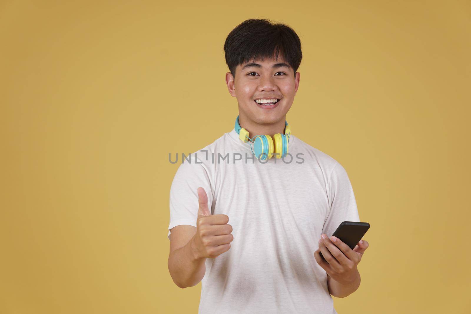 portrait of happy joyful cheerful young asian man wearing headphones listening to music on smartphone and dancing isolated on yellow studio background