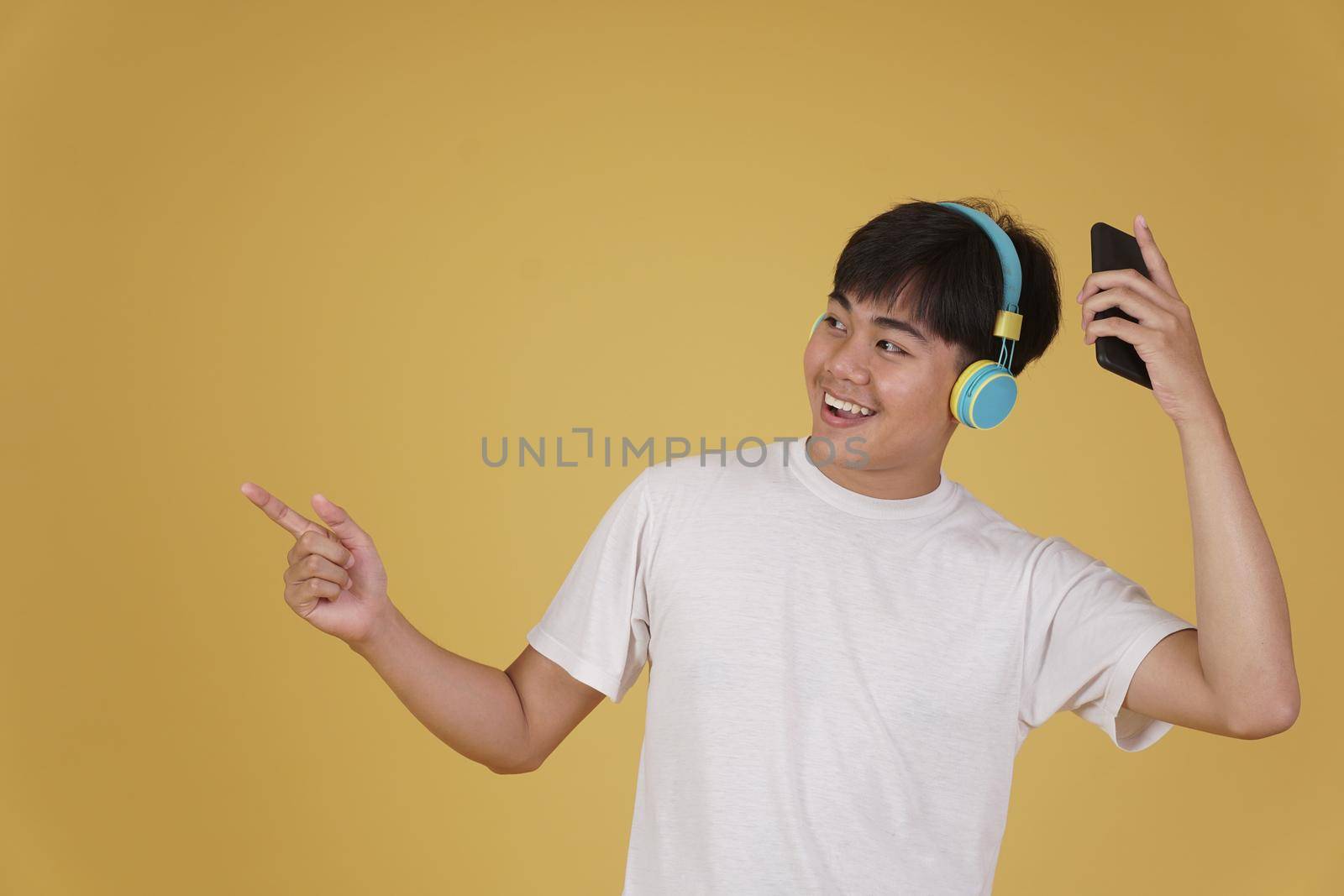 portrait of happy joyful cheerful young asian man wearing headphones listening to music on smartphone and dancing isolated on yellow studio background
