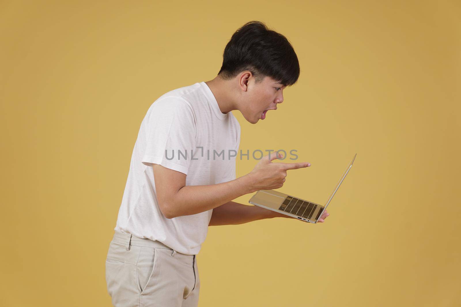 aggressive angry upset furious young asian man scolding screaming yelling on laptop computer isolated on yellow studio background