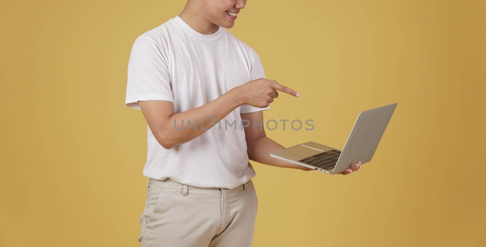 happy glad rejoicing cheerful young asian man dressed casually holding pointing finger at laptop computer screen isolated on yellow studio background