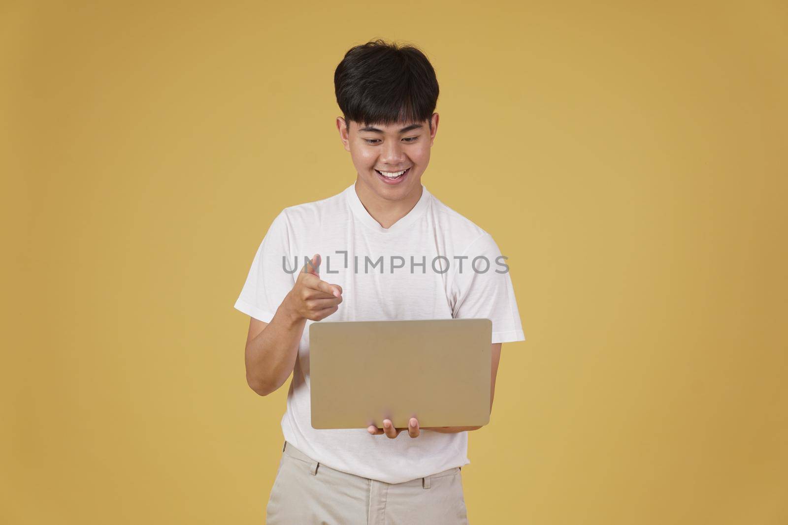 happy glad rejoicing cheerful young asian man dressed casually holding pointing finger at laptop computer screen isolated on yellow studio background