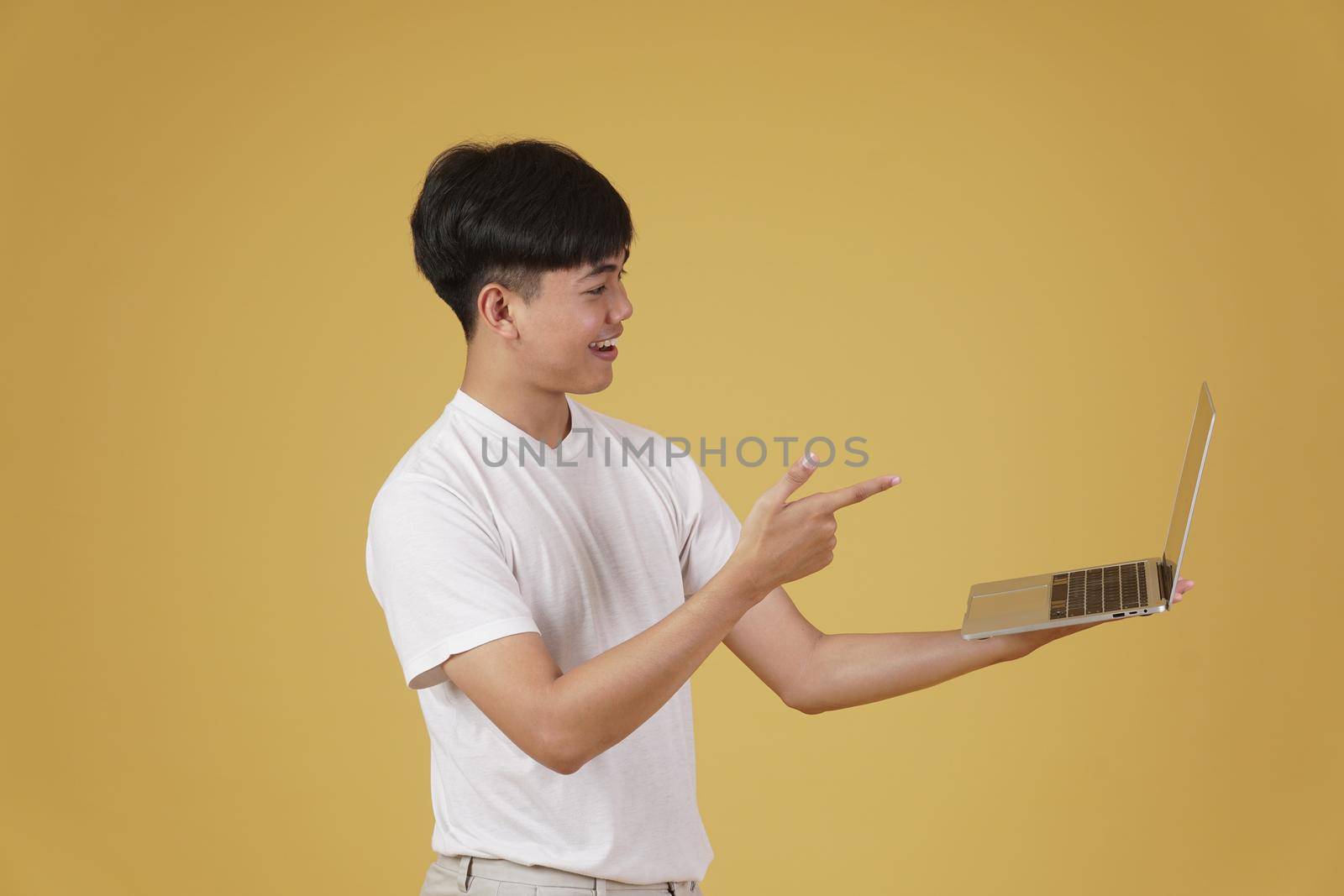 happy glad rejoicing cheerful young asian man dressed casually holding pointing finger at laptop computer screen isolated on yellow studio background