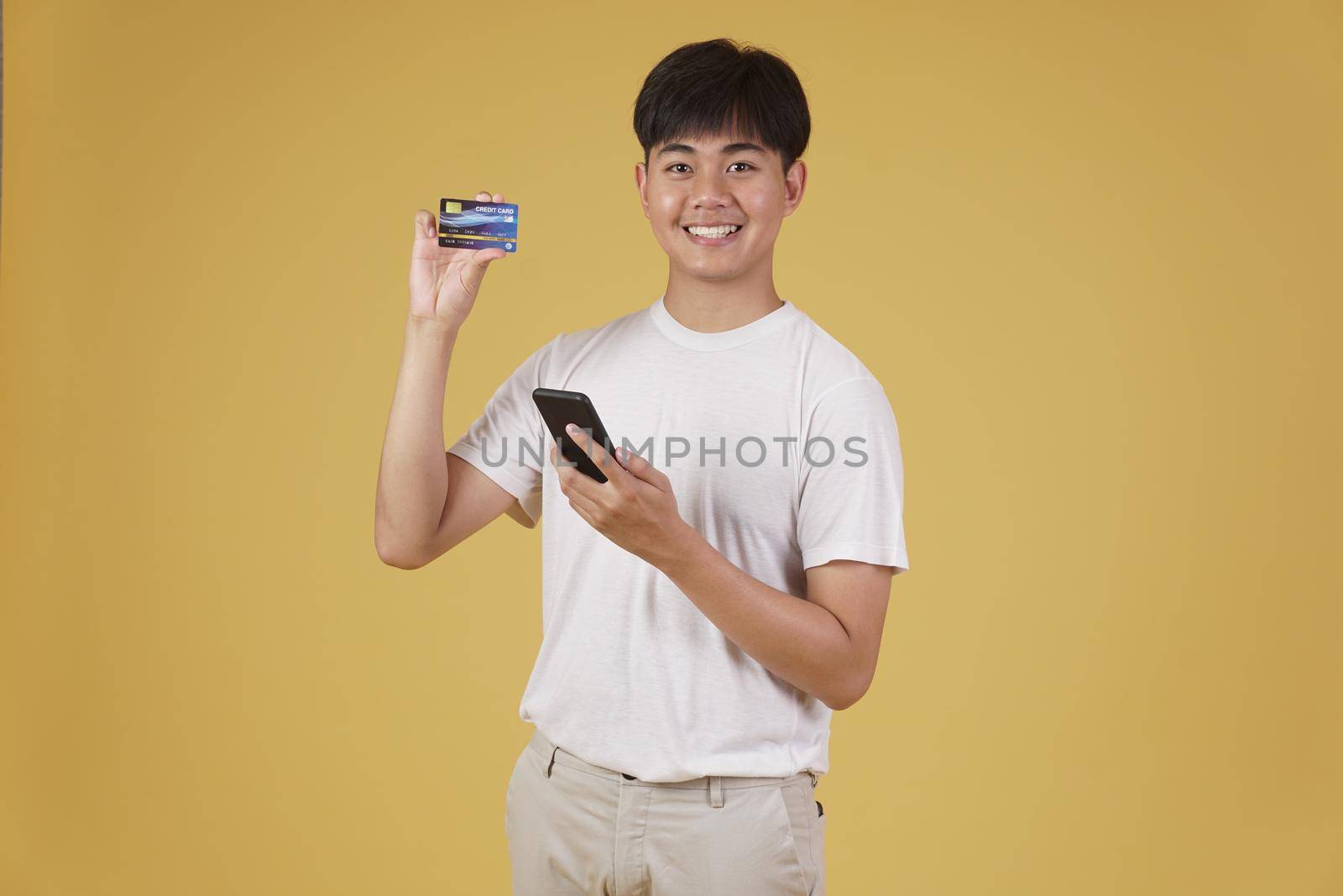 portrait of happy young asian man dressed casually holding smartphone and credit card for shopping online isolated on yellow studio background