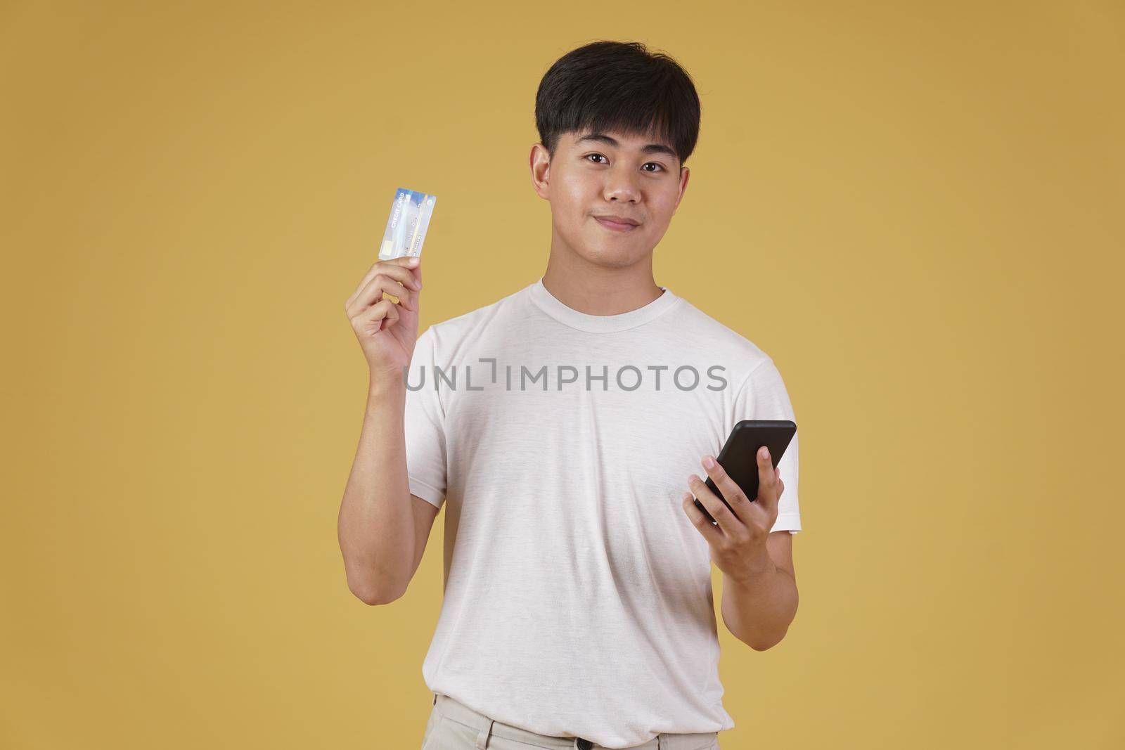 portrait of happy young asian man dressed casually holding smartphone and credit card for shopping online isolated on yellow studio background