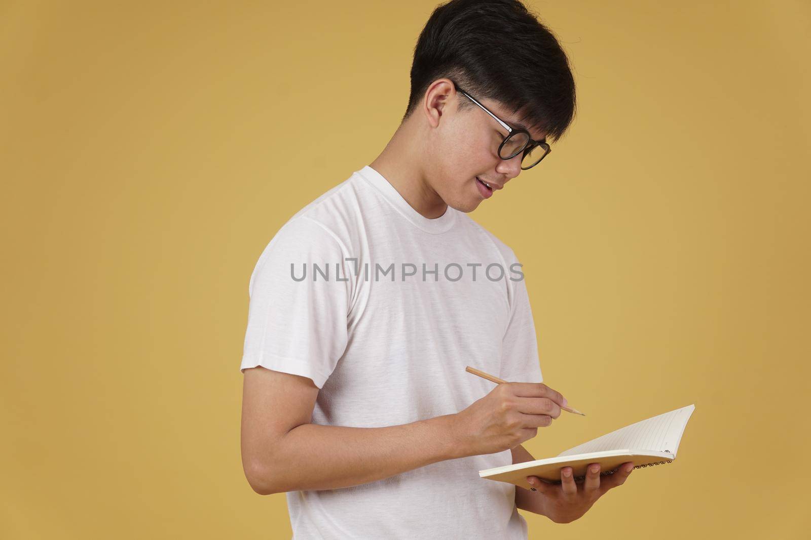 happy cheerful young asian man student dressed casually wearing eyeglasses writing note isolated on yellow studio background