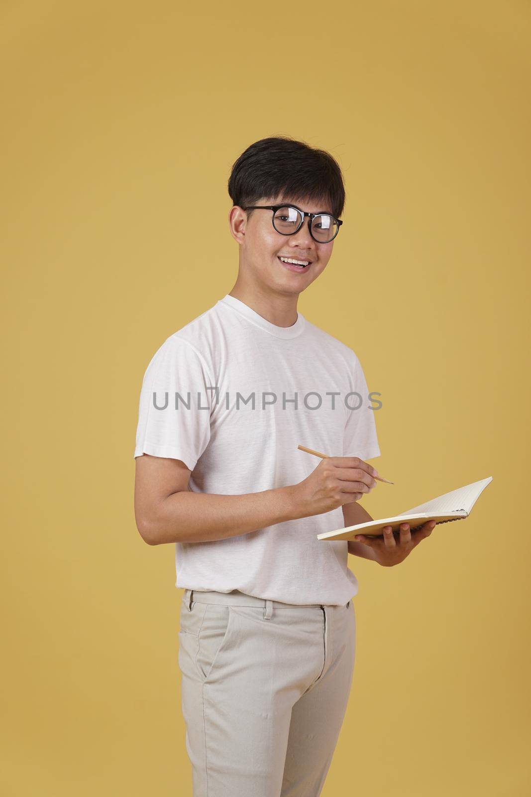 happy cheerful young asian man student dressed casually wearing eyeglasses writing note isolated on yellow studio background