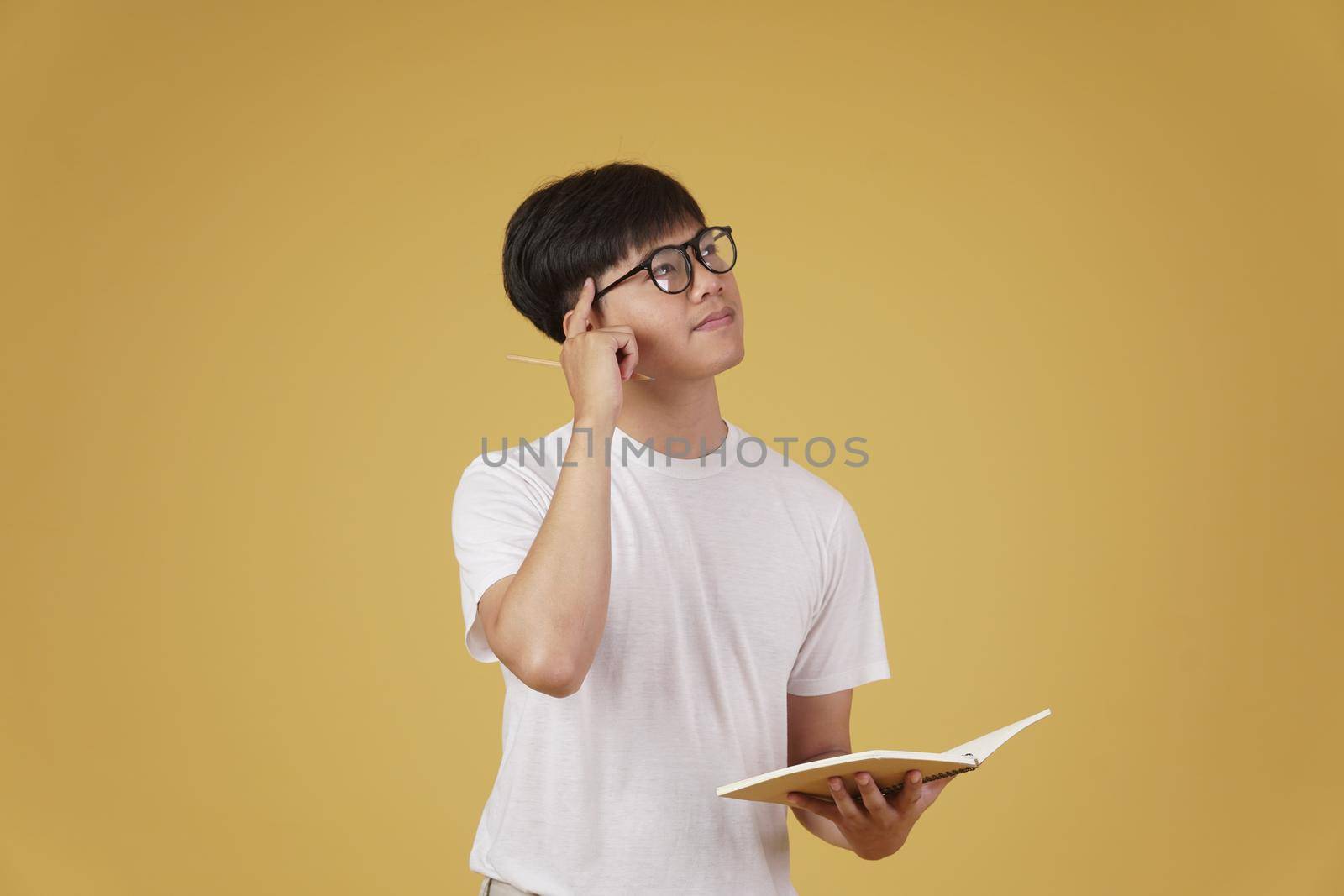 thoughtful young asian man student dressed casually wearing eyeglasses holding notebook thinking isolated on yellow studio background