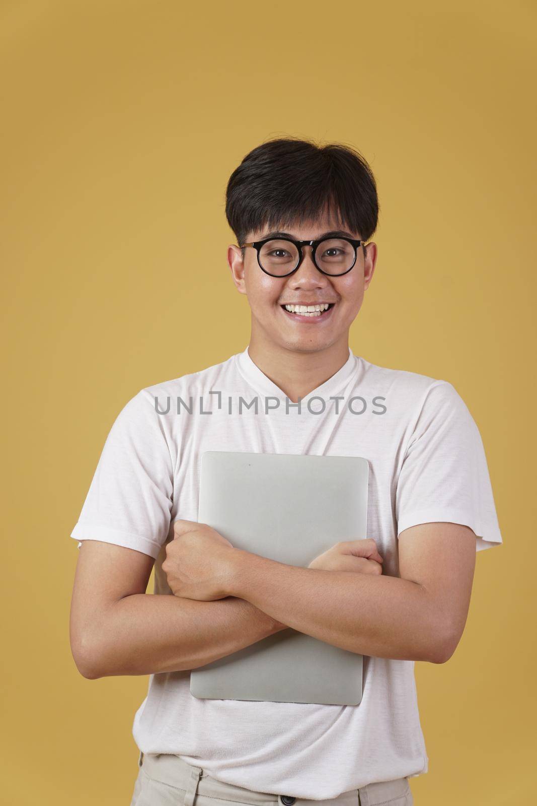 happy cheerful young asian man nerd student dressed casually wearing eyeglasses holding laptop computer isolated on yellow studio background