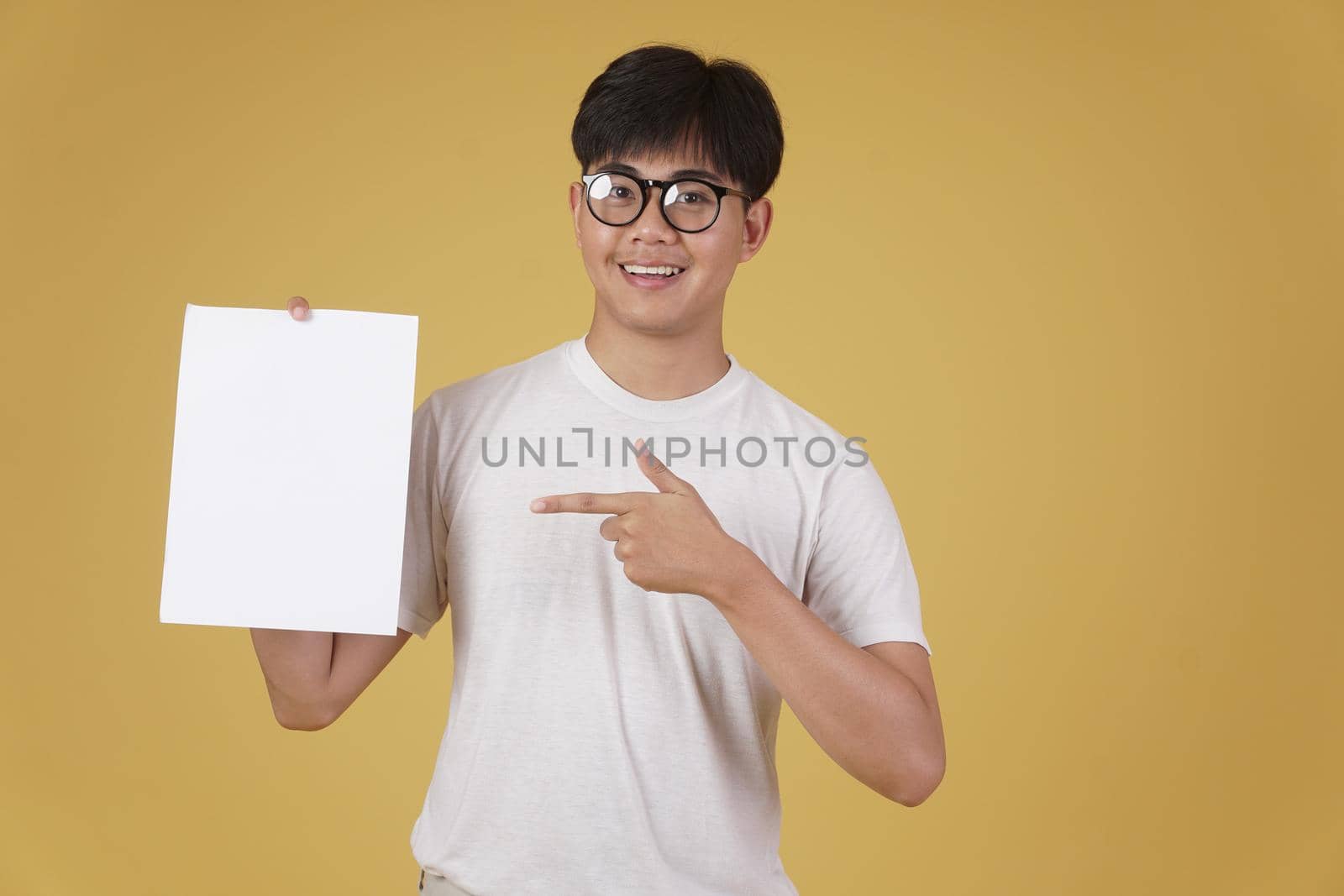 portrait of smiling happy cheerful young asian man dressed casually showing blank empty placard paper isolated on yellow studio background.