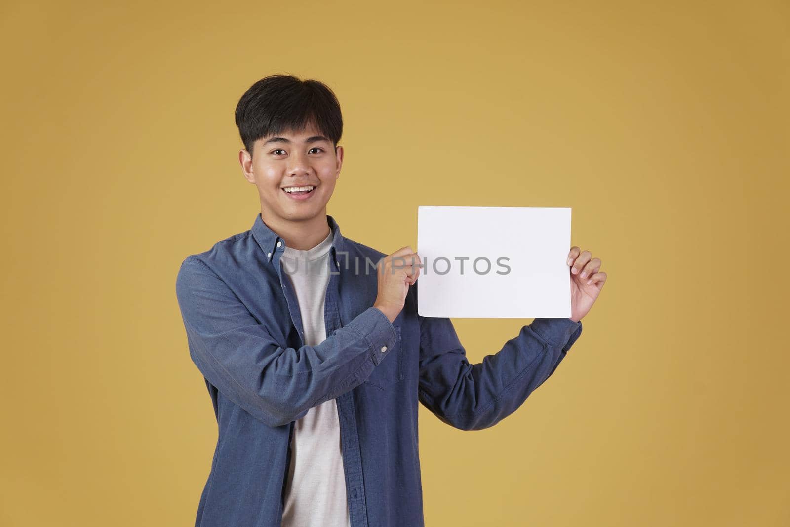 portrait of smiling happy cheerful young asian man dressed casually showing blank empty placard paper isolated on yellow studio background.