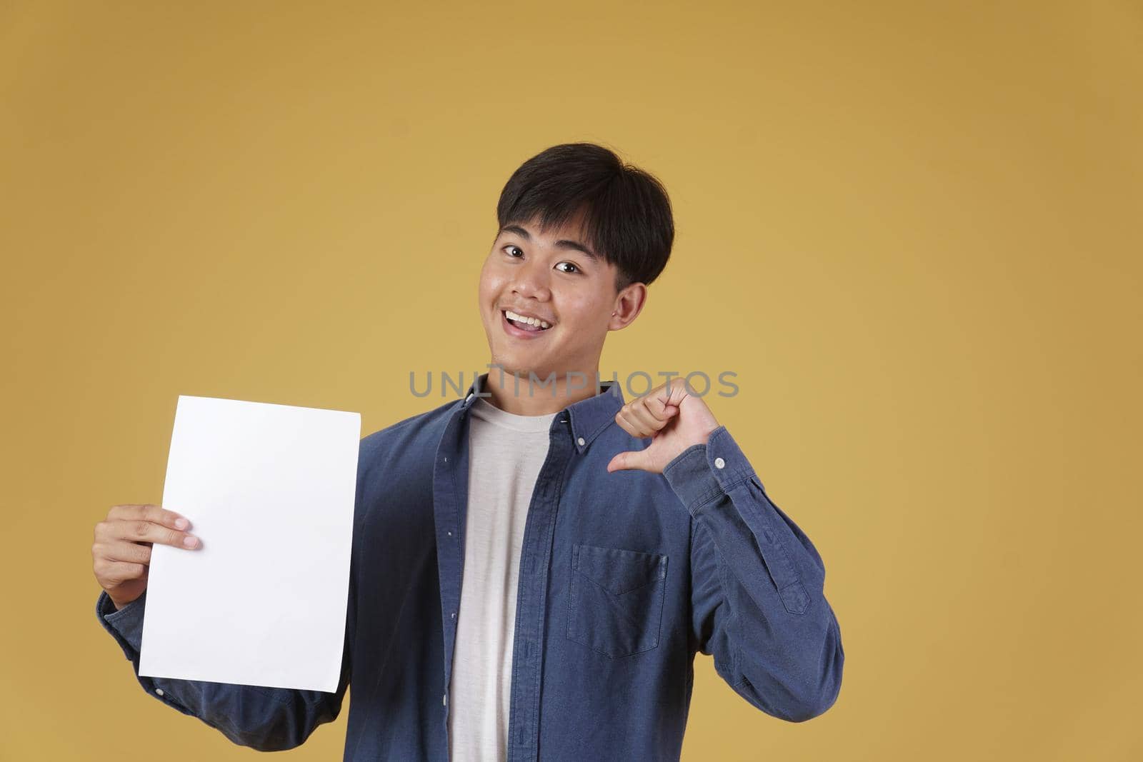 portrait of smiling happy cheerful young asian man dressed casually showing blank empty placard paper pointing thumb to himself isolated on yellow studio background.