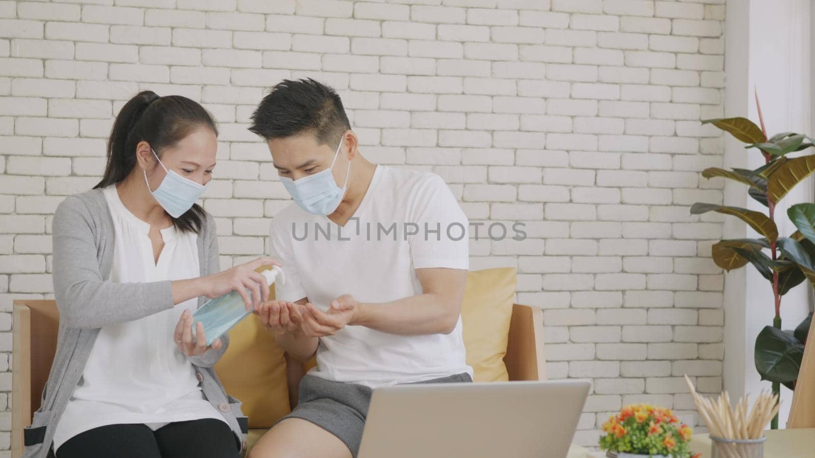 Happy Asian beautiful family couple husband and wife laughing sitting on sofa in the living room working with laptop computer at home. Woman brings alcohol gel applying to hand man, COVID-19
