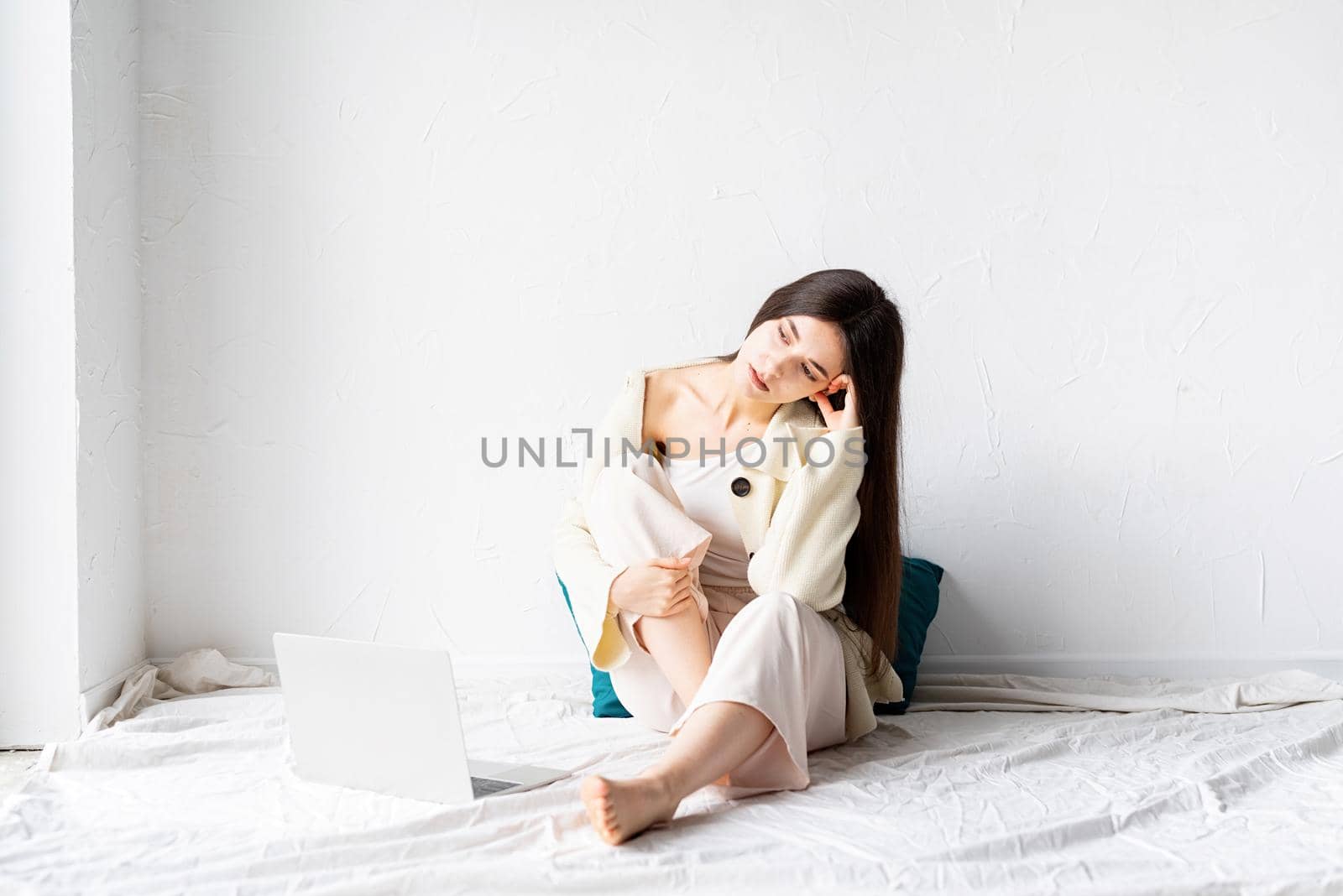 Beautiful young woman sitting on the floor and doing freelance project on laptop, looking away